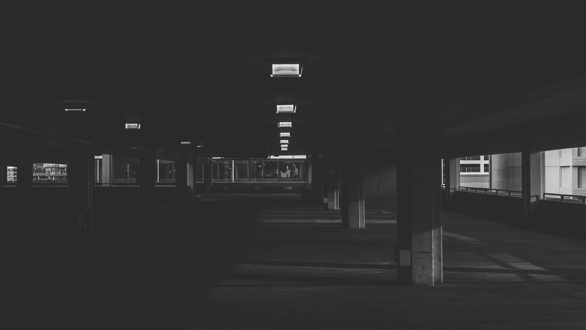 單色地下停車場 壁紙 — 一條長長嘅昏暗地下通道，有混凝土柱同遠處發光嘅出口標誌，投射出鮮明嘅陰影。