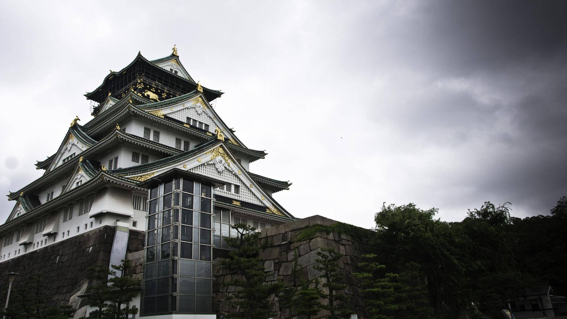單色大阪城 壁紙 — 一座雄偉的日本城堡，白色牆壁和金色點綴，矗立在戲劇性的灰色天空下，周圍環繞著石牆和綠樹。