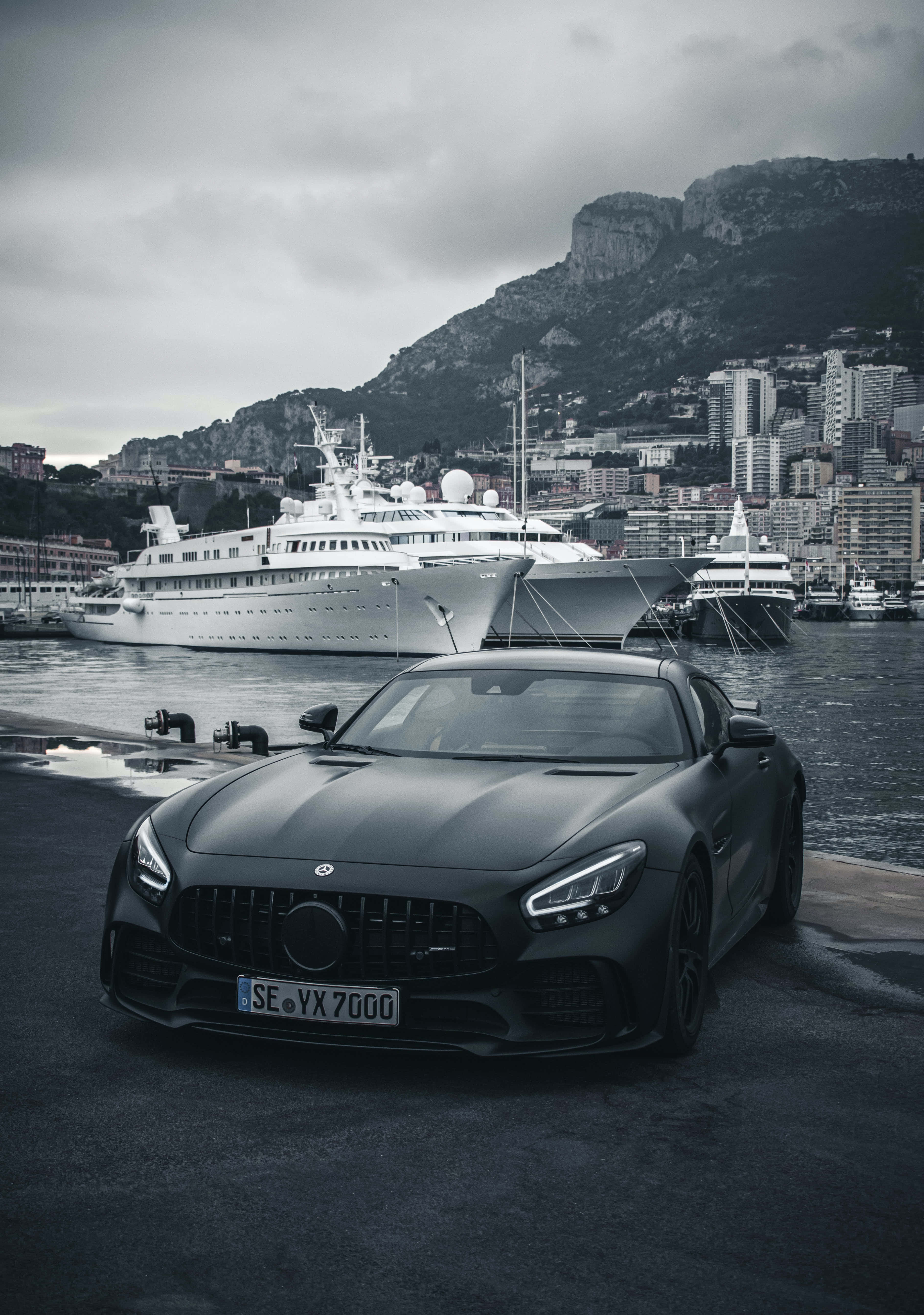 單色梅賽德斯-奔馳 AMG GT R 背景 壁紙 — 一輛啞光黑色跑車停泊在港口碼頭，背景是陰天，有豪華遊艇和山區沿海城市。