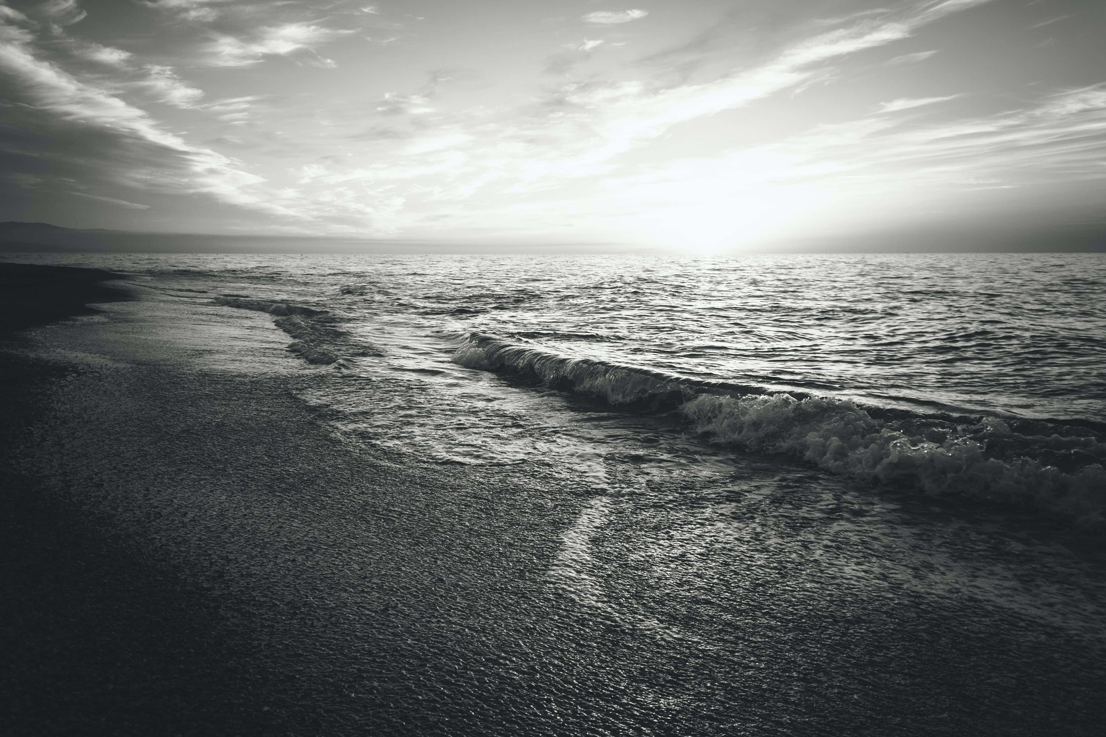 Black And White Ocean 桌布 壁紙 — 單色海灘場景，柔和的海浪在明亮的天空下拍打濕沙。