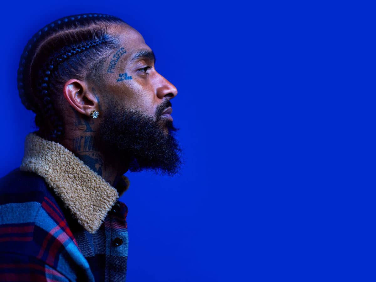 單色藍色Crips Nipsey Hussle 壁紙 — 一個辮子頭髮和紋身的男人，在純藍色背景上若有所思地凝視著。