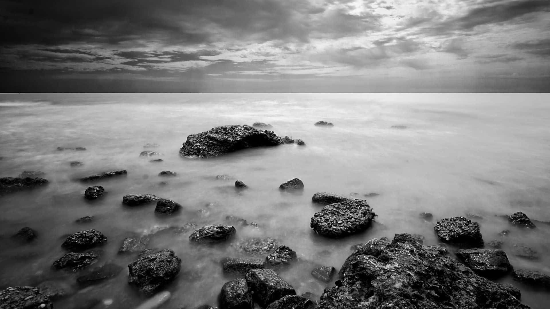 Dark Landscape 桌布 壁紙 — 單色長曝光海景，平滑的水流繞過有紋理的岩石，在戲劇性的多雲天空下。