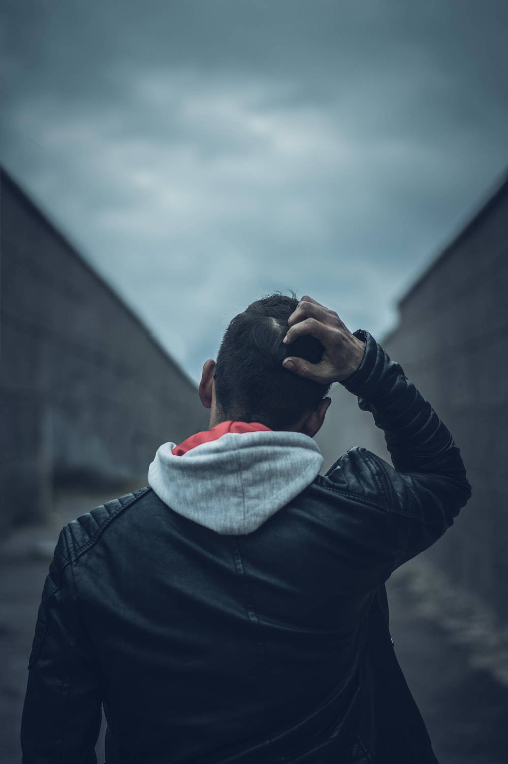 單身男孩 頭部姿勢 壁紙 — 一個穿著黑色皮褸的男人背對鏡頭，手放在頭上，在陰沉天空下被高聳的混凝土牆框住。