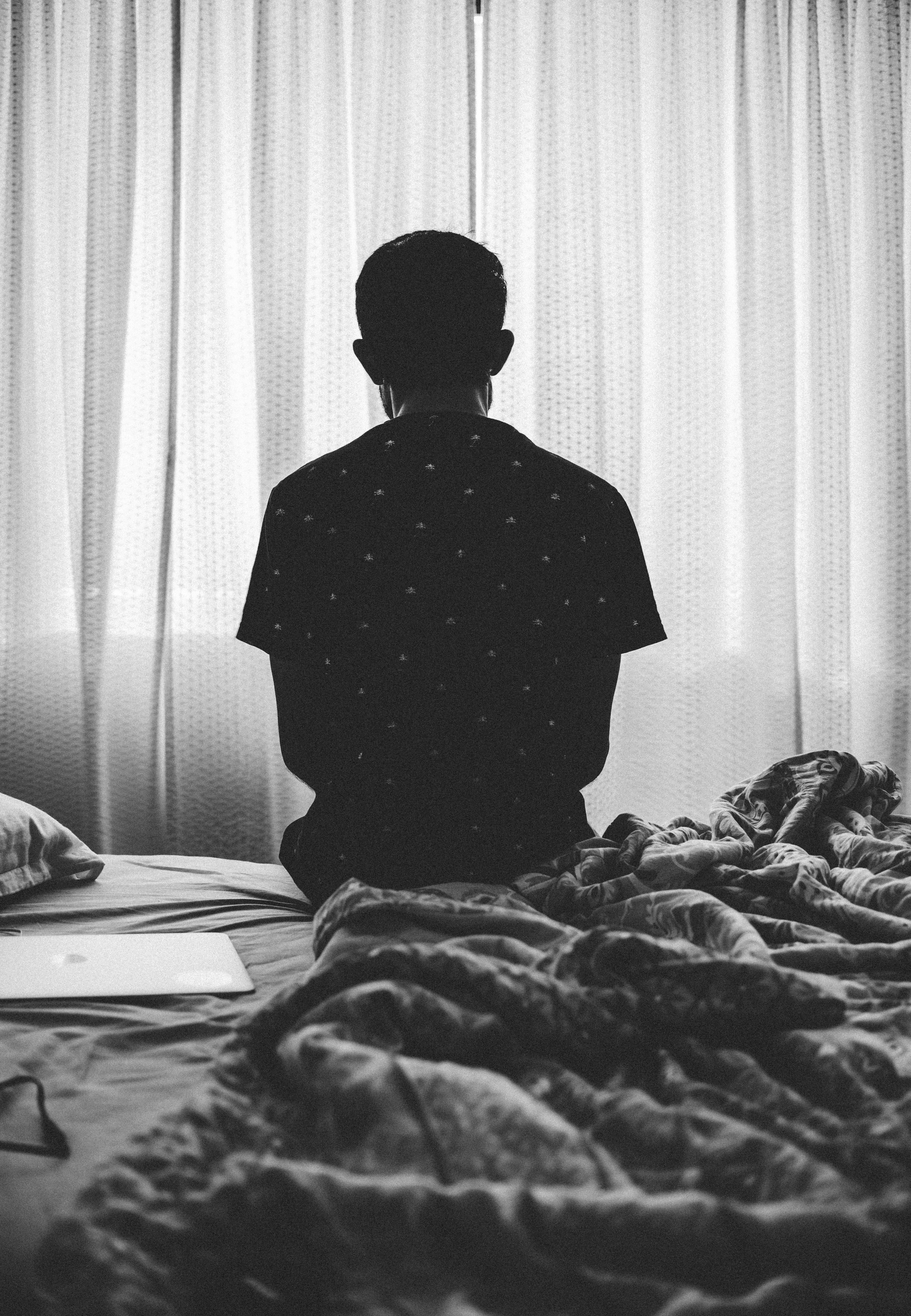單身男孩坐床上 壁紙 — 一個人坐在皺巴巴的床上，面向透光窗簾，在單色房間中，柔和自然光線透入。