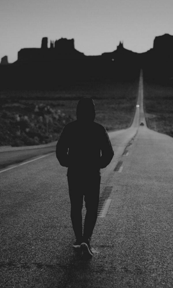 單身男孩獨自行走於長路 壁紙 — 一個孤獨的戴帽人影在黃昏時沿著一條漫長的沙漠道路走向遠處的岩石地貌。