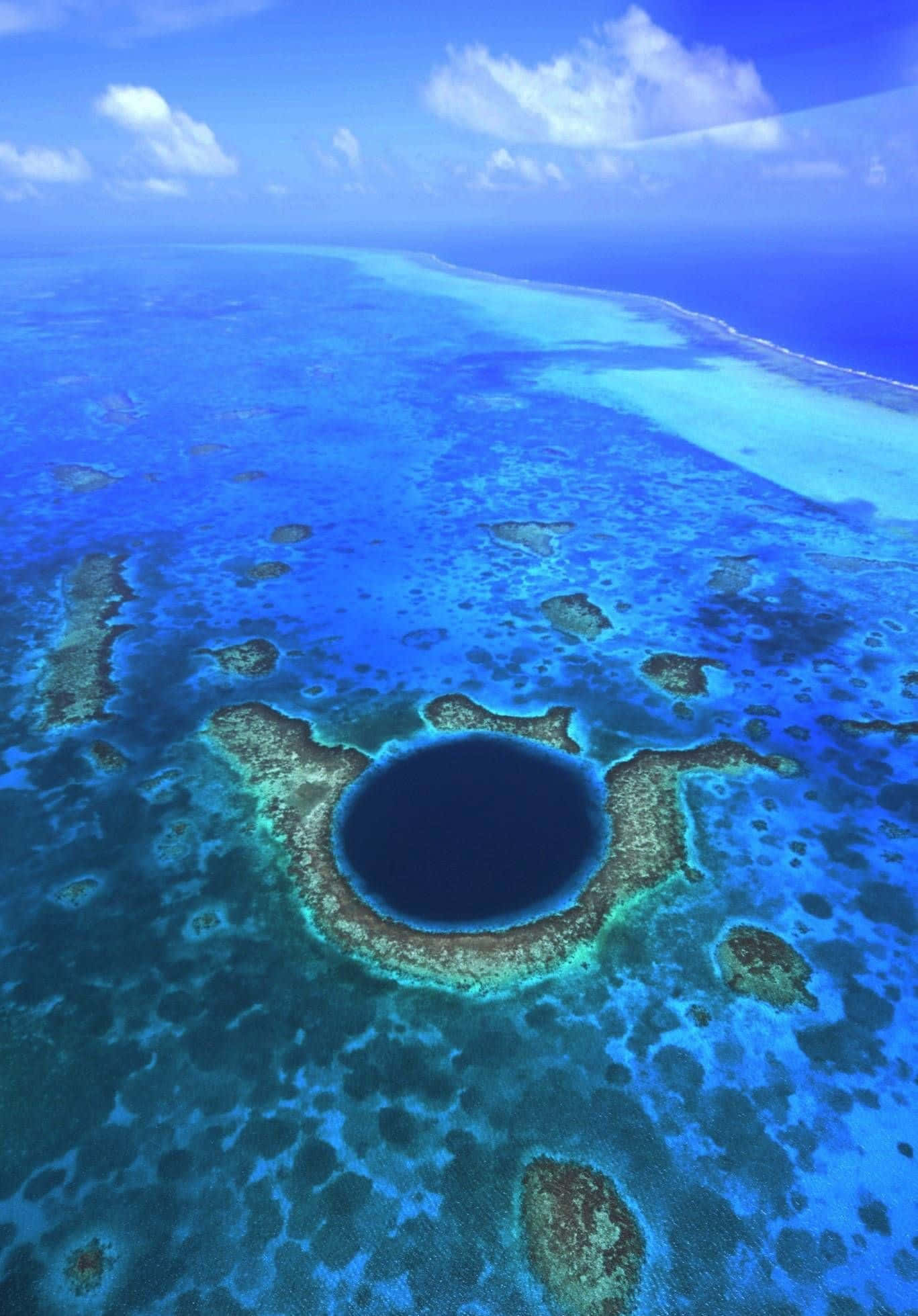Blue Hole 桌布 壁紙 — 喺明亮天空下，一個被充滿活力嘅珊瑚礁同藍綠色海洋包圍嘅深圓形藍色沉洞嘅鳥瞰圖。