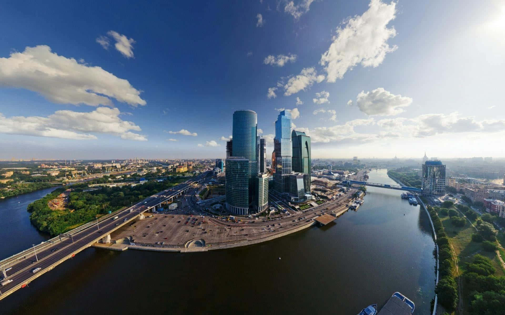 Russia 桌布 壁紙 — 喺明亮藍天同零星白雲下，現代玻璃摩天大廈沿住一條寬闊河流排列，河上有橋樑同翠綠公園嘅航拍景觀。