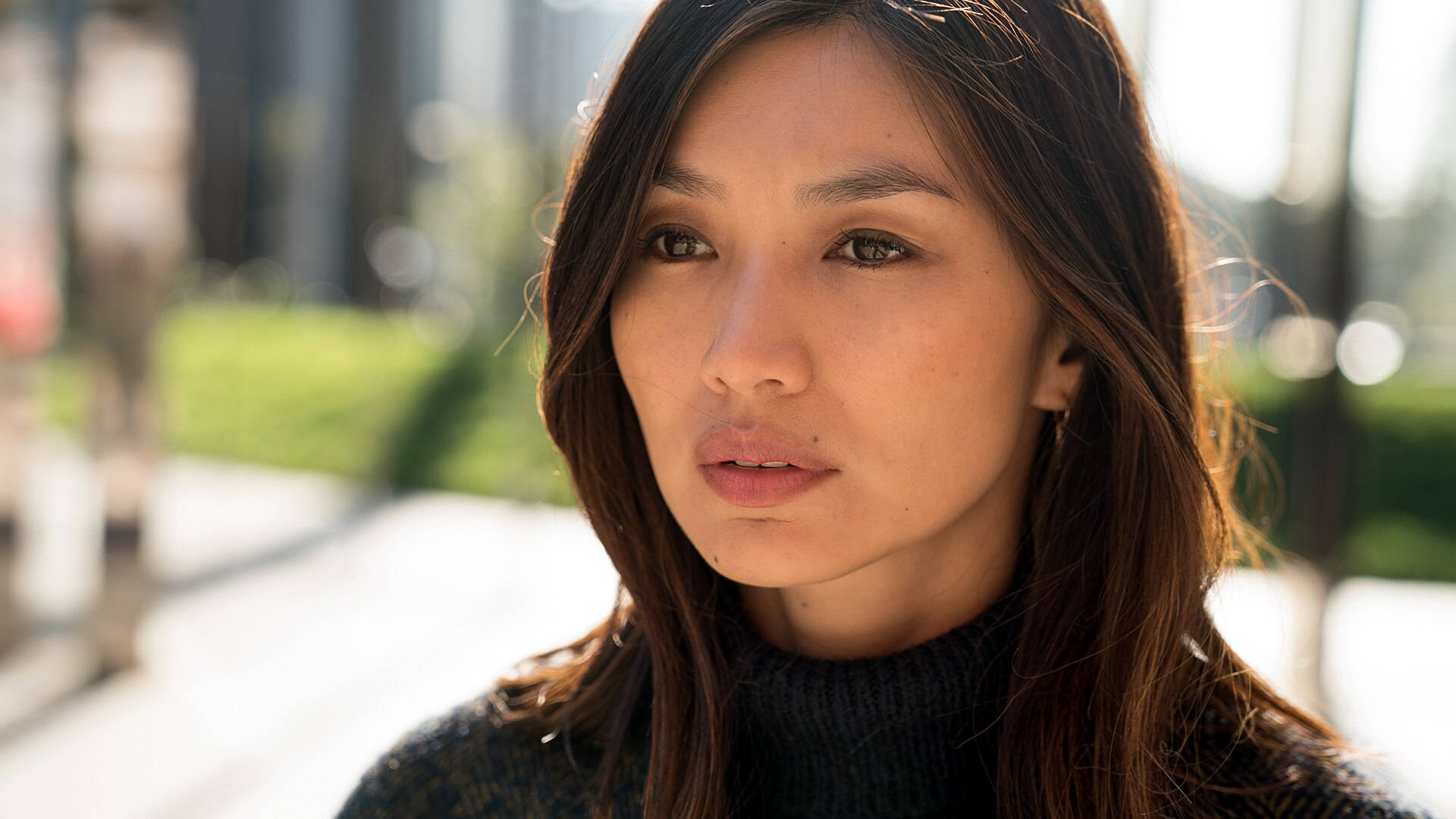 嘉瑪·陳 憂慮凝視 壁紙 — 一位深色頭髮、穿著黑色高領毛衣的女性在晴朗的日子裡若有所思地望著戶外，身後是模糊的城市綠化。