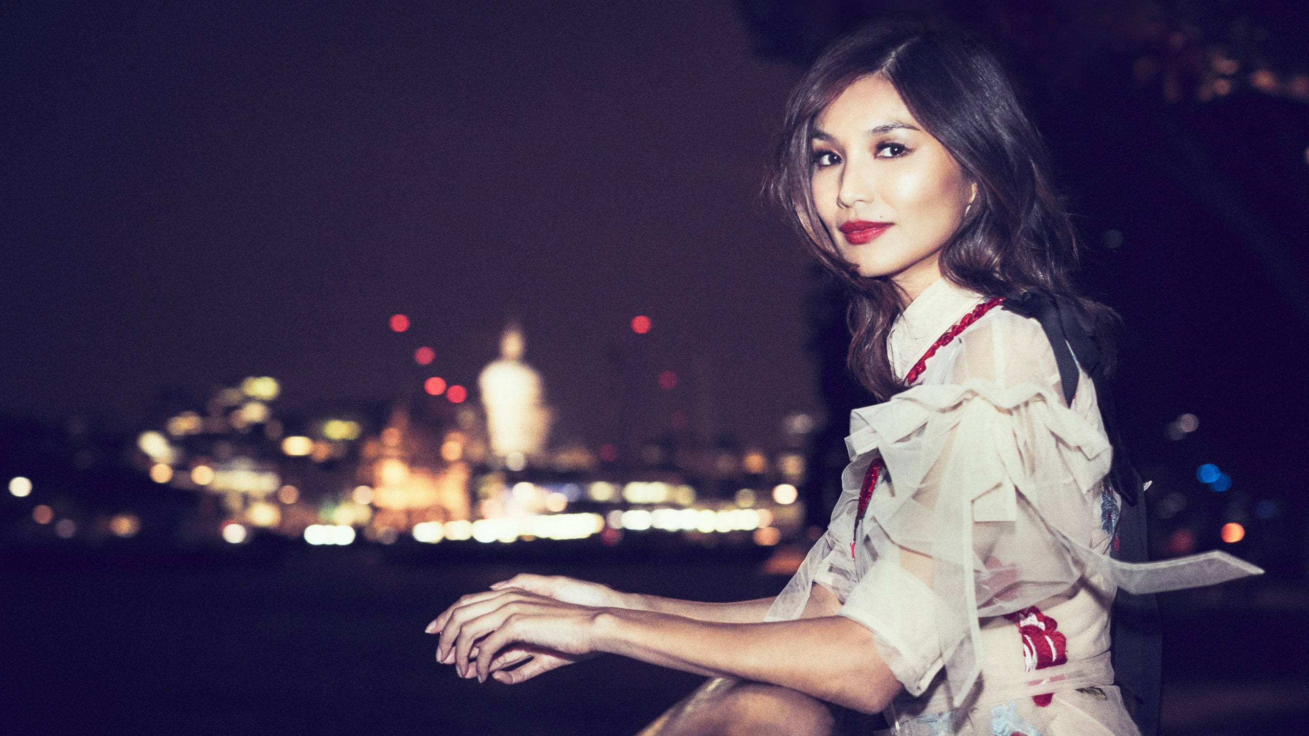 嘉瑪·陳夜景海邊 壁紙 — 一位身穿荷葉邊白色連衣裙的女性在夜晚坐在水邊，身後是散景的城市景觀閃爍著光芒。