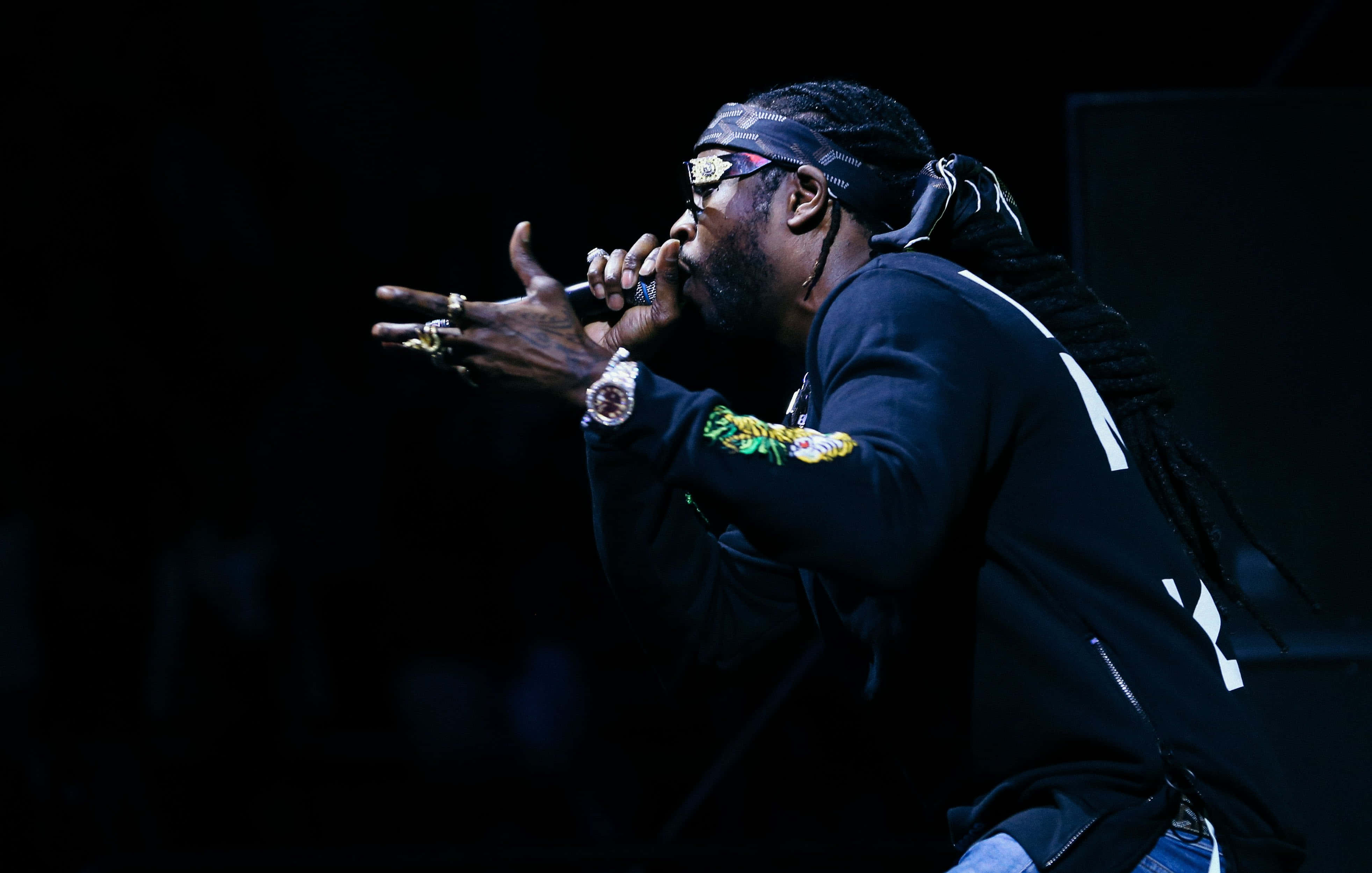 嘻哈巨星2 Chainz為鏡頭擺姿 壁紙 — 一位梳著辮子頭、戴著太陽眼鏡的饒舌歌手，在戲劇性的舞台燈光下，對著麥克風充滿激情地表演。