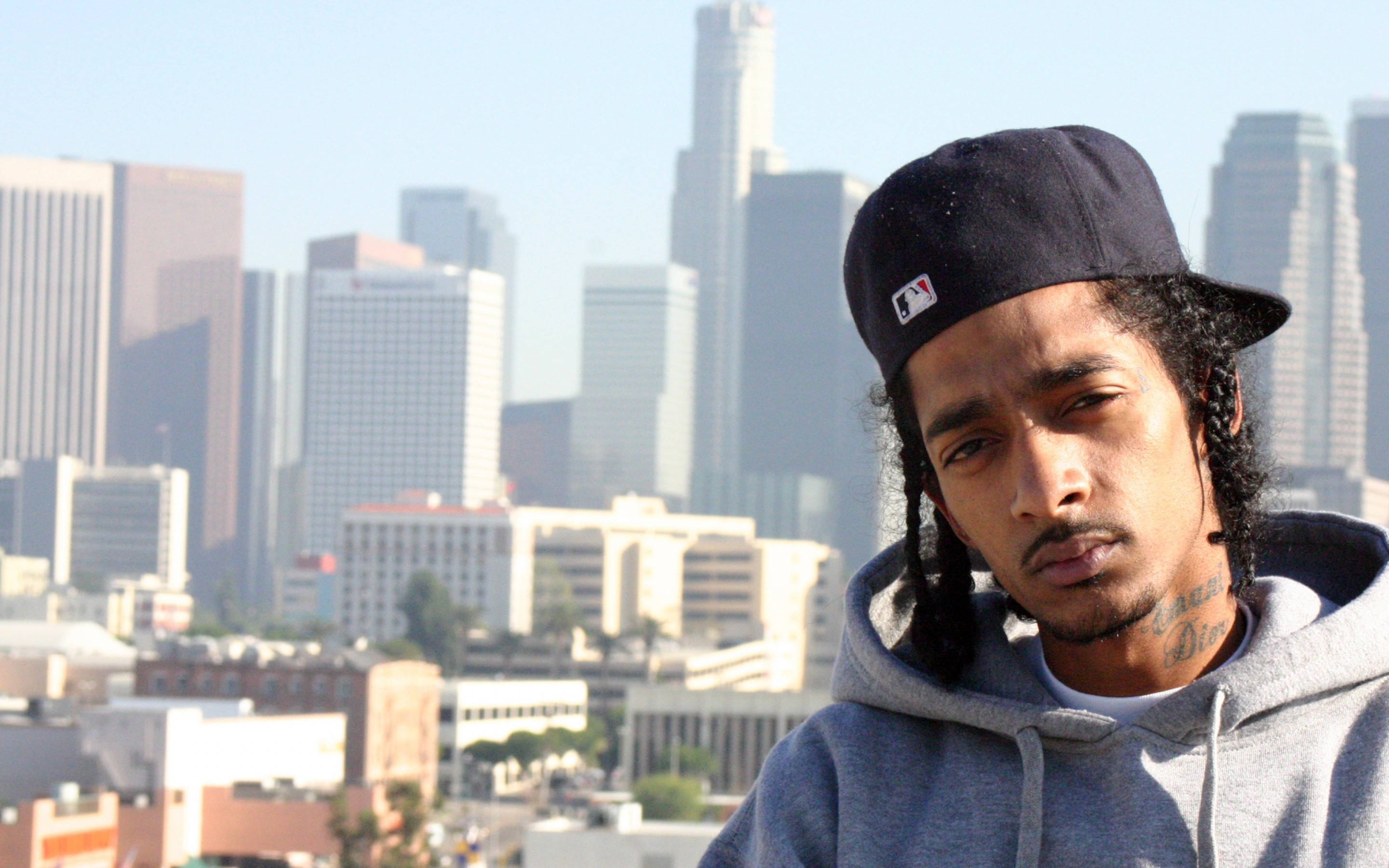 嘻哈界傳奇人物 Nipsey Hussle 壁紙 — 一名年輕男子編著辮子，戴著黑色帽子，在明亮的日光下凝視著朦朧的洛杉磯天際線。