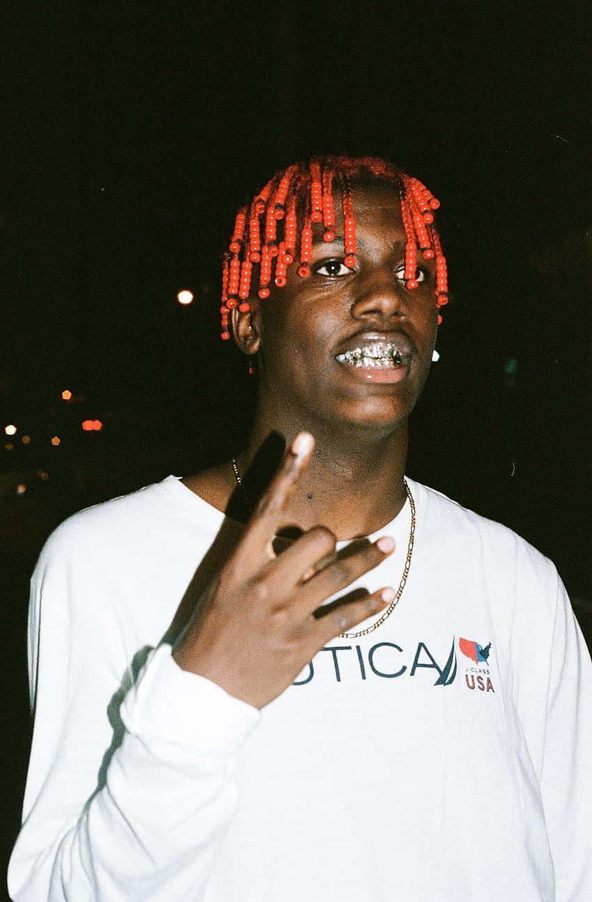 嘻哈藝人Lil Yachty 壁紙 — 一位年輕男子，擁有鮮紅色辮子頭髮和銀色牙套，在黑暗的夜晚城市燈光背景下微笑並做出手勢。