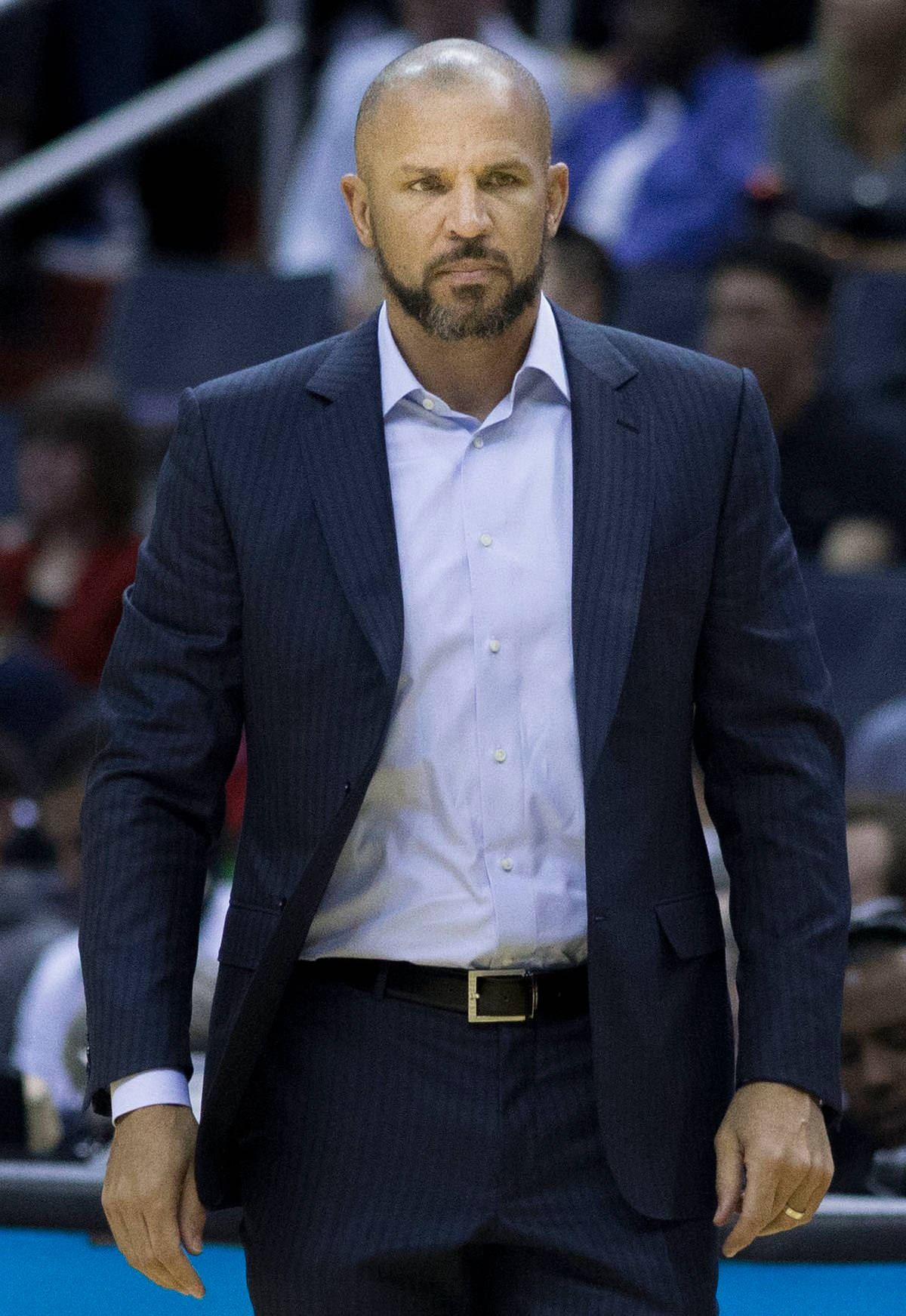 嚴肅的NBA教練賈森·基德 壁紙 — 一位光頭鬍鬚的籃球教練穿著深色細條紋西裝，在比賽期間專注地站在球場上。