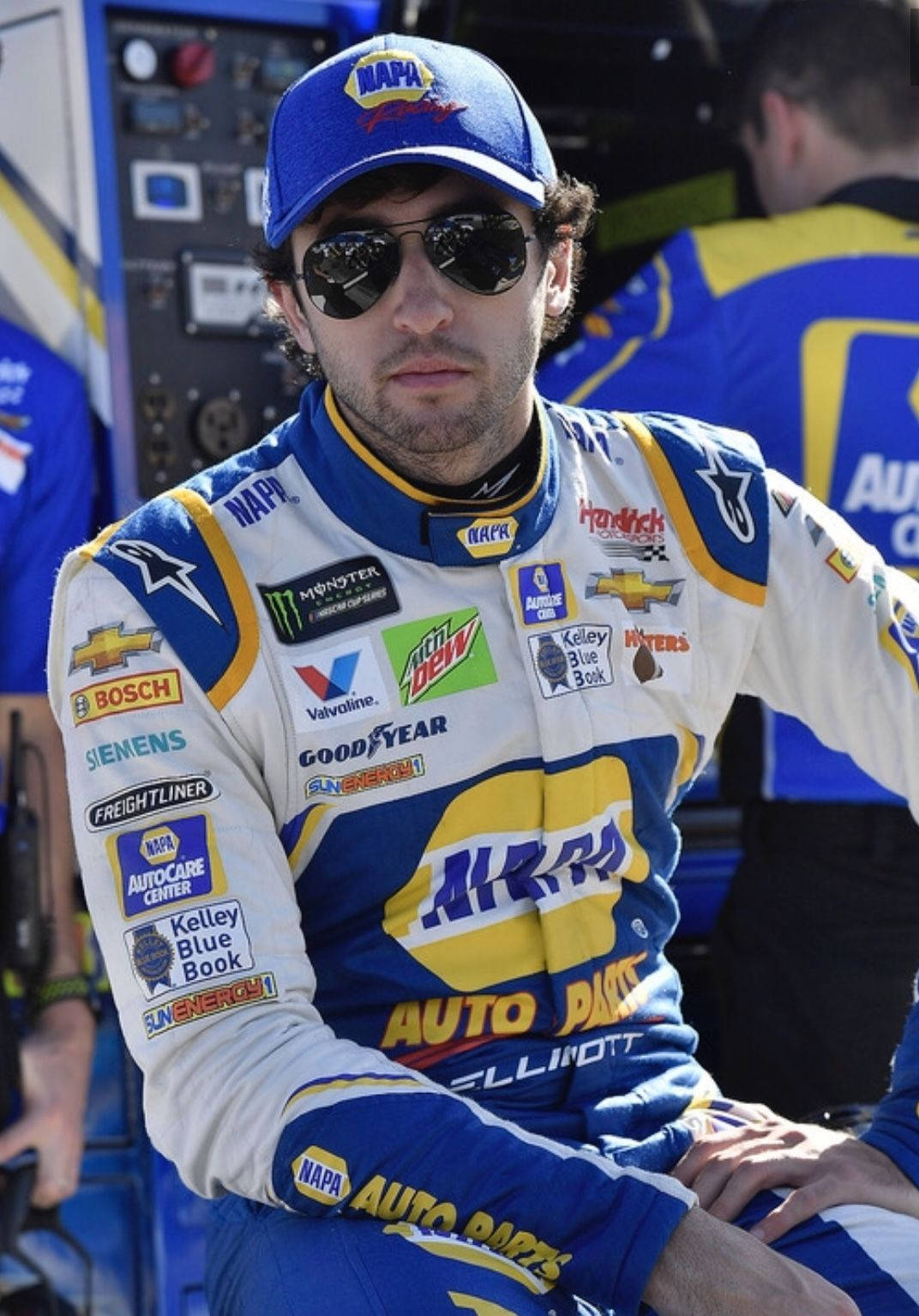 身穿藍黃賽車服的NASCAR車手，戴著太陽眼鏡和帽子。