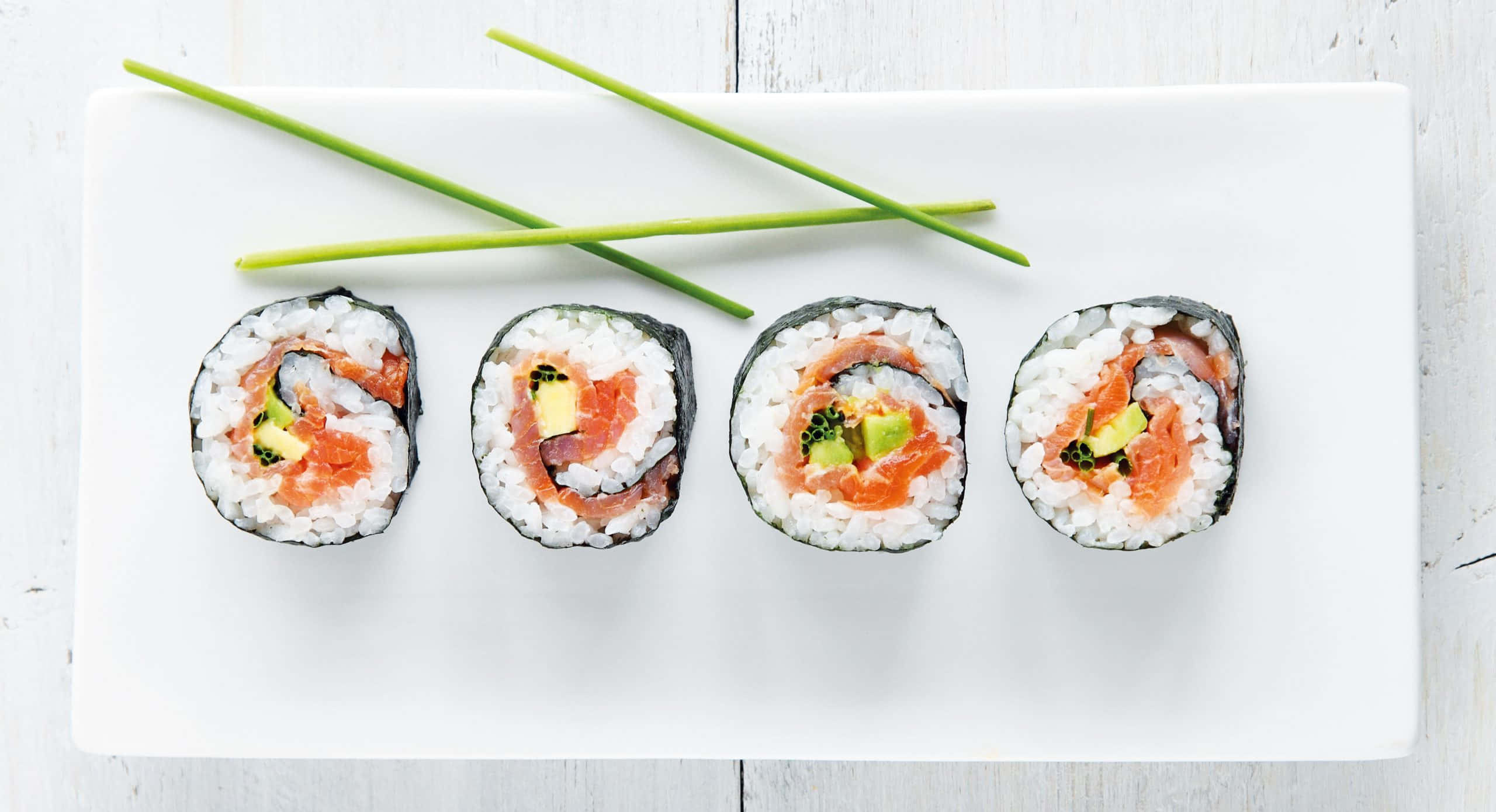 Sushi 桌布 壁紙 — 四件三文魚牛油果壽司卷整齊地排列在白色碟上，並以交叉的綠色細香蔥點綴。