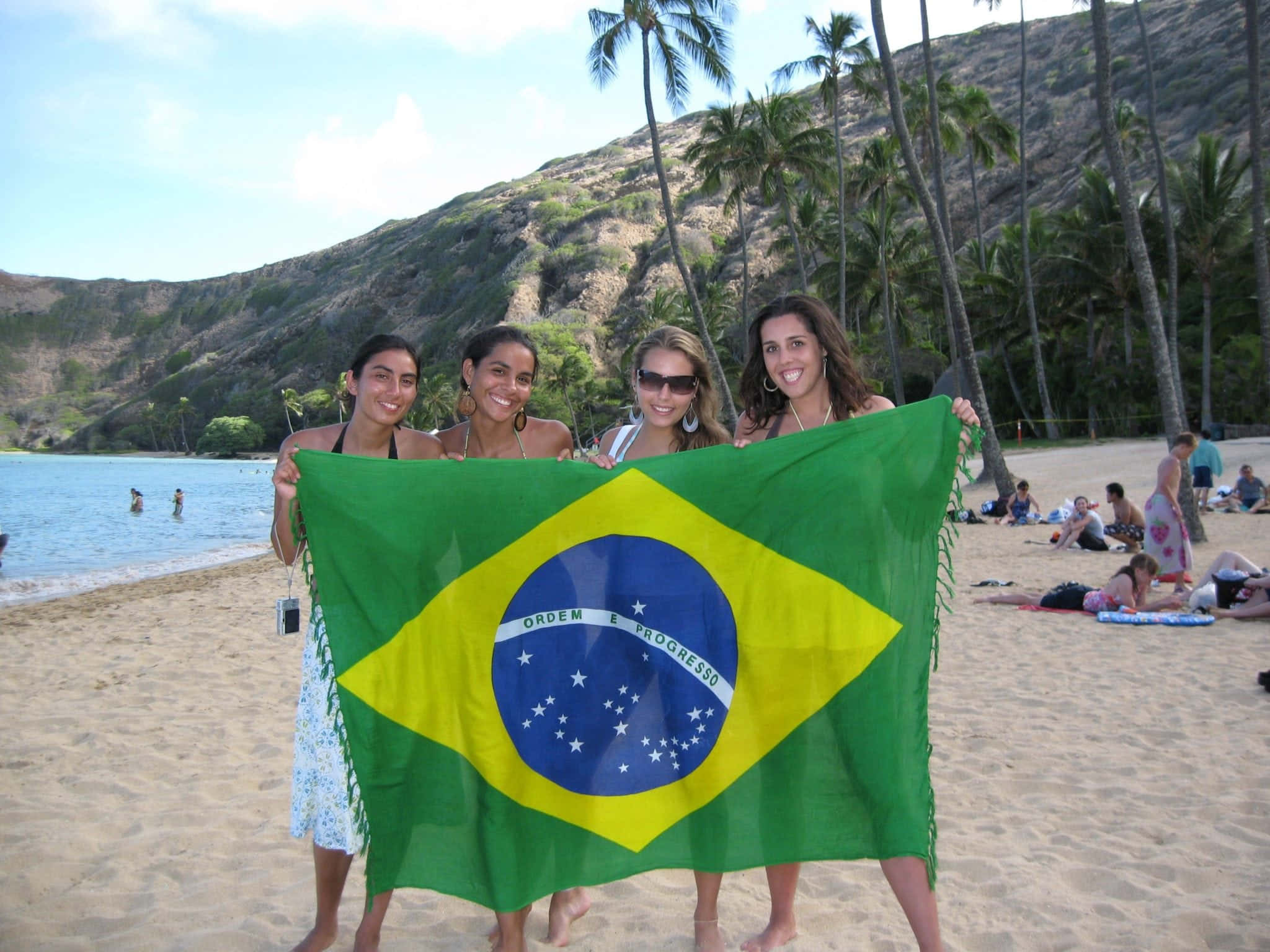 Brazilian Beach 桌布 壁紙 — 四位微笑的女性在陽光明媚的海灘上手持一面大型巴西國旗，身後有棕櫚樹和岩石山丘。