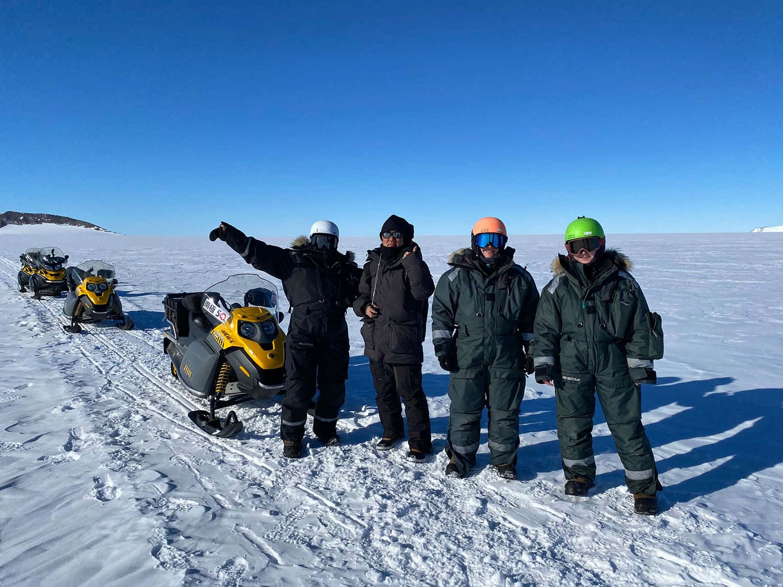 Antarctica 桌布 壁紙 — 四位戴著鮮艷頭盔的雪地摩托車手，在晴朗的藍天下，在一片廣闊的冰凍平原上，自豪地站在他們的黃色機器旁。