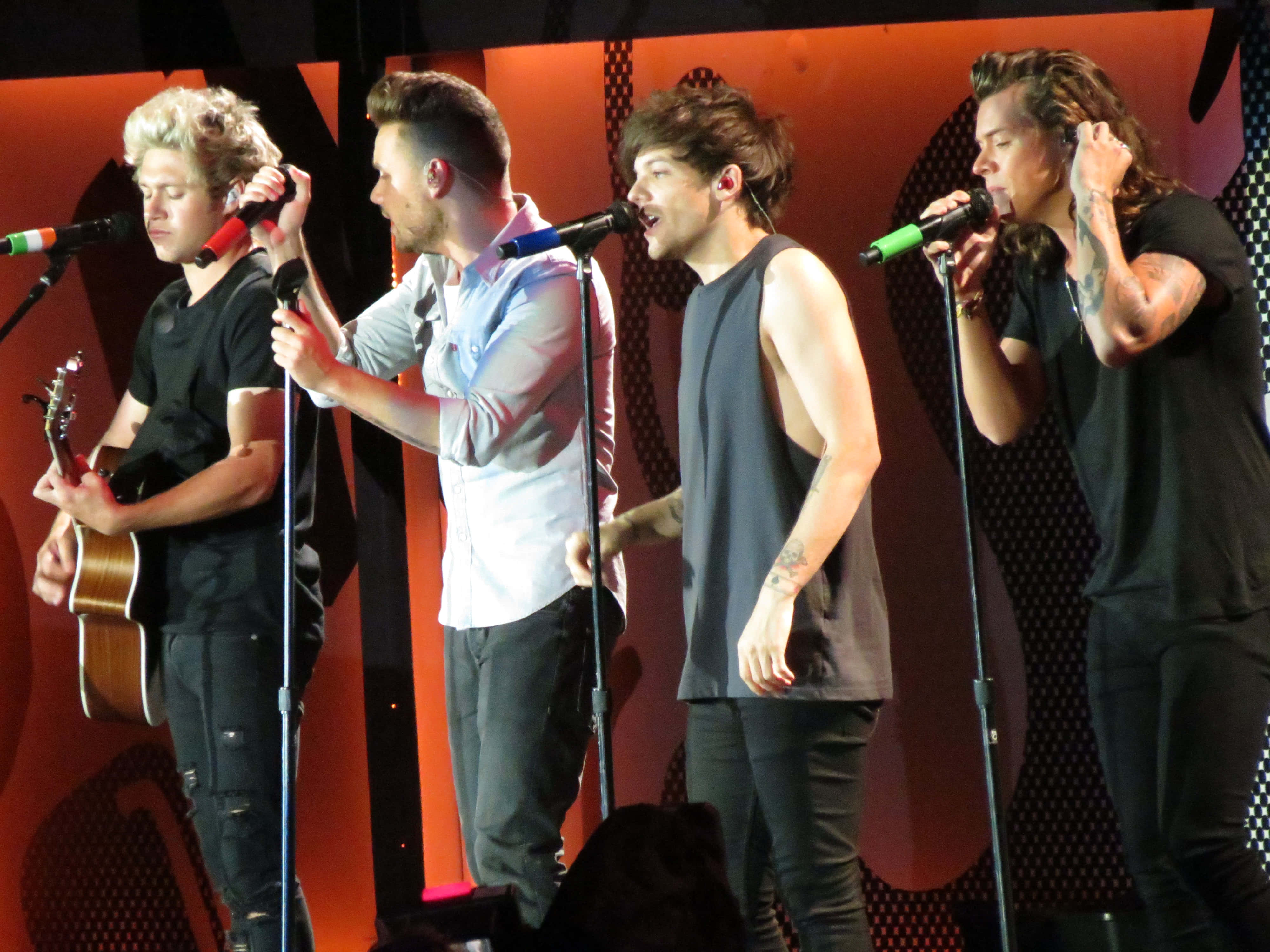 One Direction 桌布 壁紙 — 四位男性樂隊成員在溫暖的橙色燈光下，手持麥克風和結他，在舞台上充滿活力地表演。