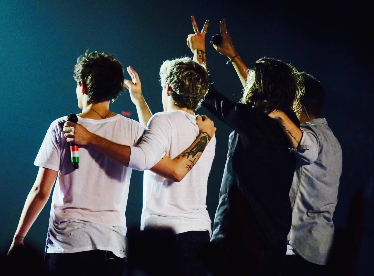 One Direction Aesthetic 桌布 壁紙 — 四位男性表演者在舞台上背對鏡頭，在藍色舞台燈光下舉起和平手勢。