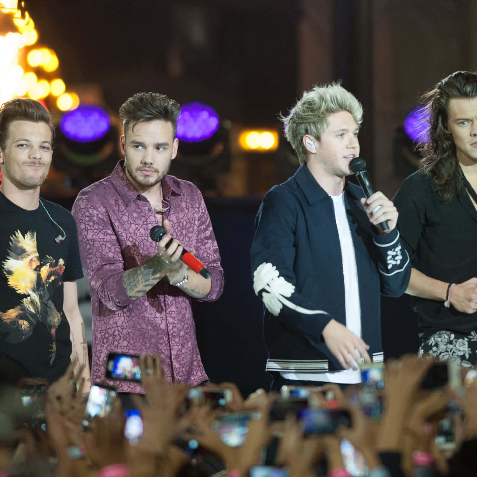 One Direction 桌布 壁紙 — 四位男音樂家在夜晚燈光明亮的舞台上表演，下方粉絲舉著手機。