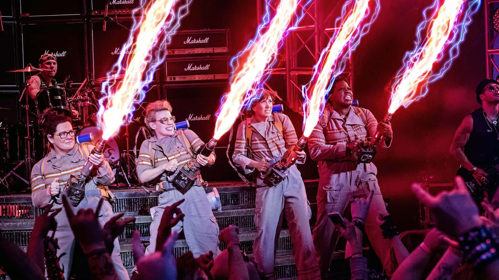 Ghostbusters 桌布 壁紙 — 四位穿著標誌性制服的捉鬼敢死隊在音樂會舞台上使用質子背包，發射出明亮的橙色和藍色能量光束。