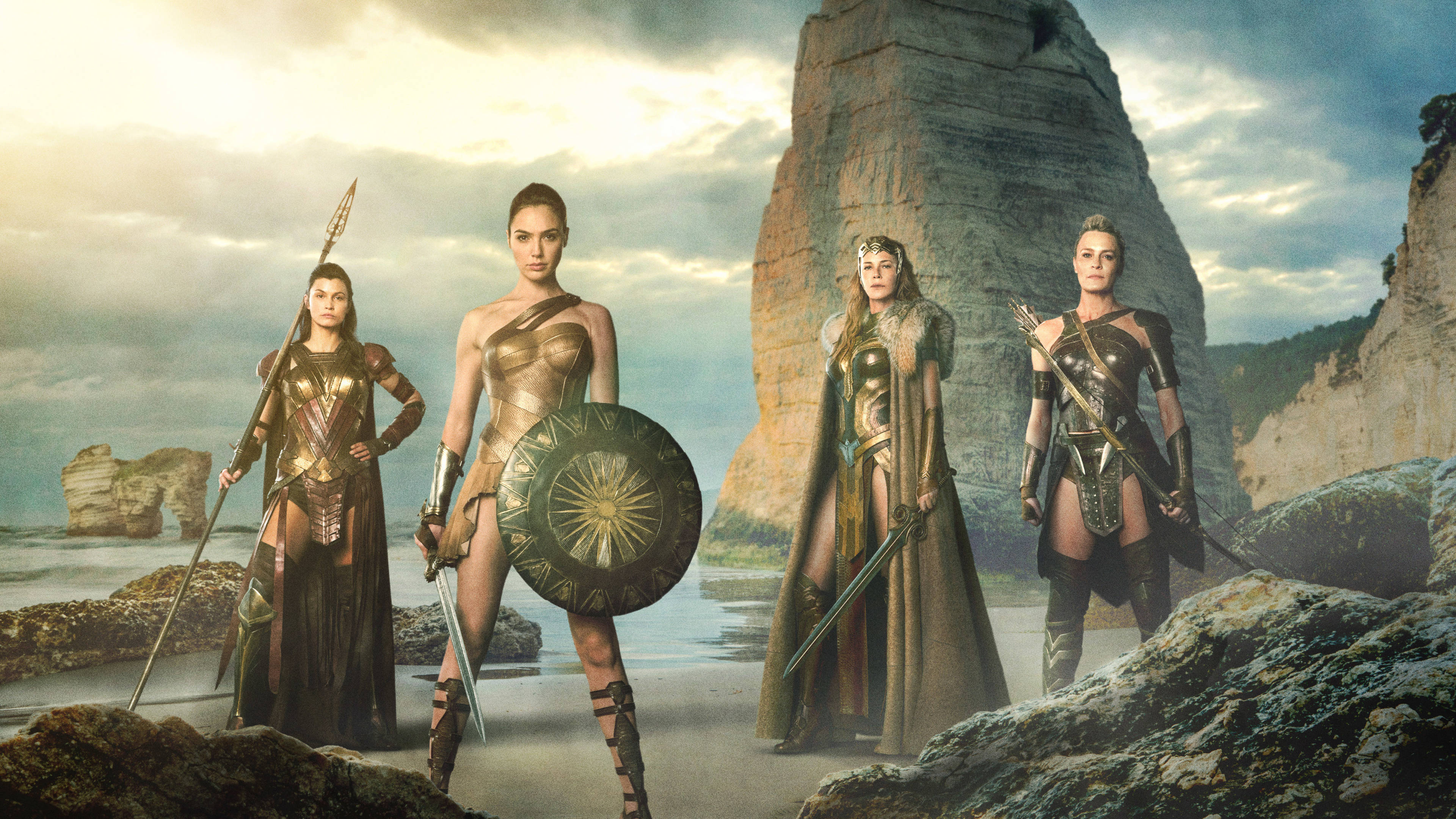 Wonder Woman unites Amazon warriors in epic fight for justice. 壁紙 — 四位身穿金色盔甲的亞馬遜戰士，在陰雲密布的天空下，挑釁地站在岩石海灘上，旁邊是高聳的懸崖。