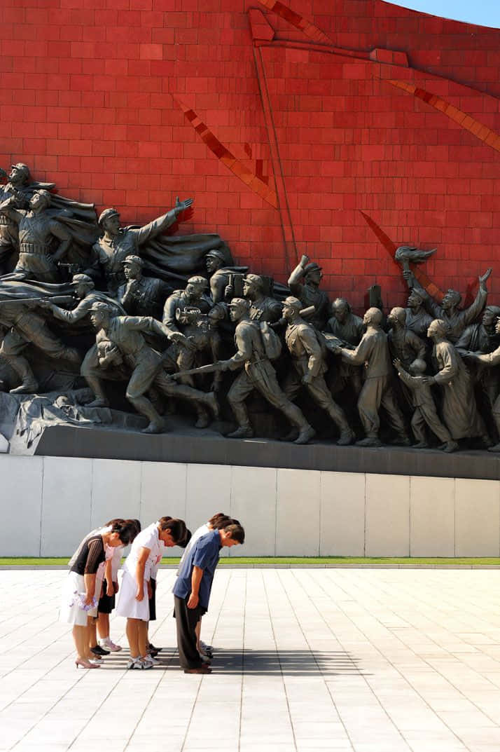 North Korea 桌布 壁紙 — 四個人恭敬地鞠躬，面前是一座大型青銅紀念碑，描繪士兵在鮮豔的紅磚牆前行動。