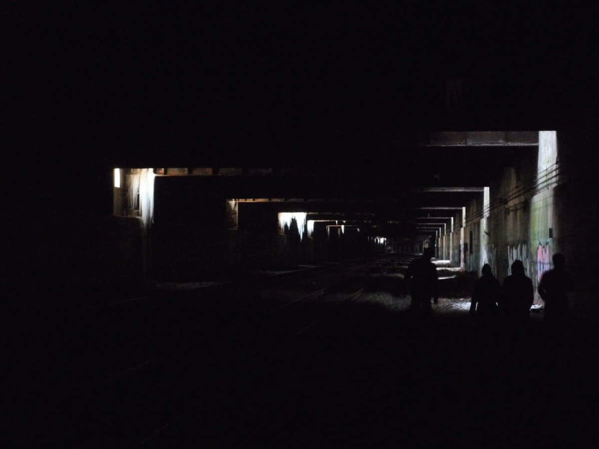 Dark Underground 桌布 壁紙 — 四個人的剪影走過一條黑暗廢棄的鐵路隧道，被遠處閃爍的燈光照亮。