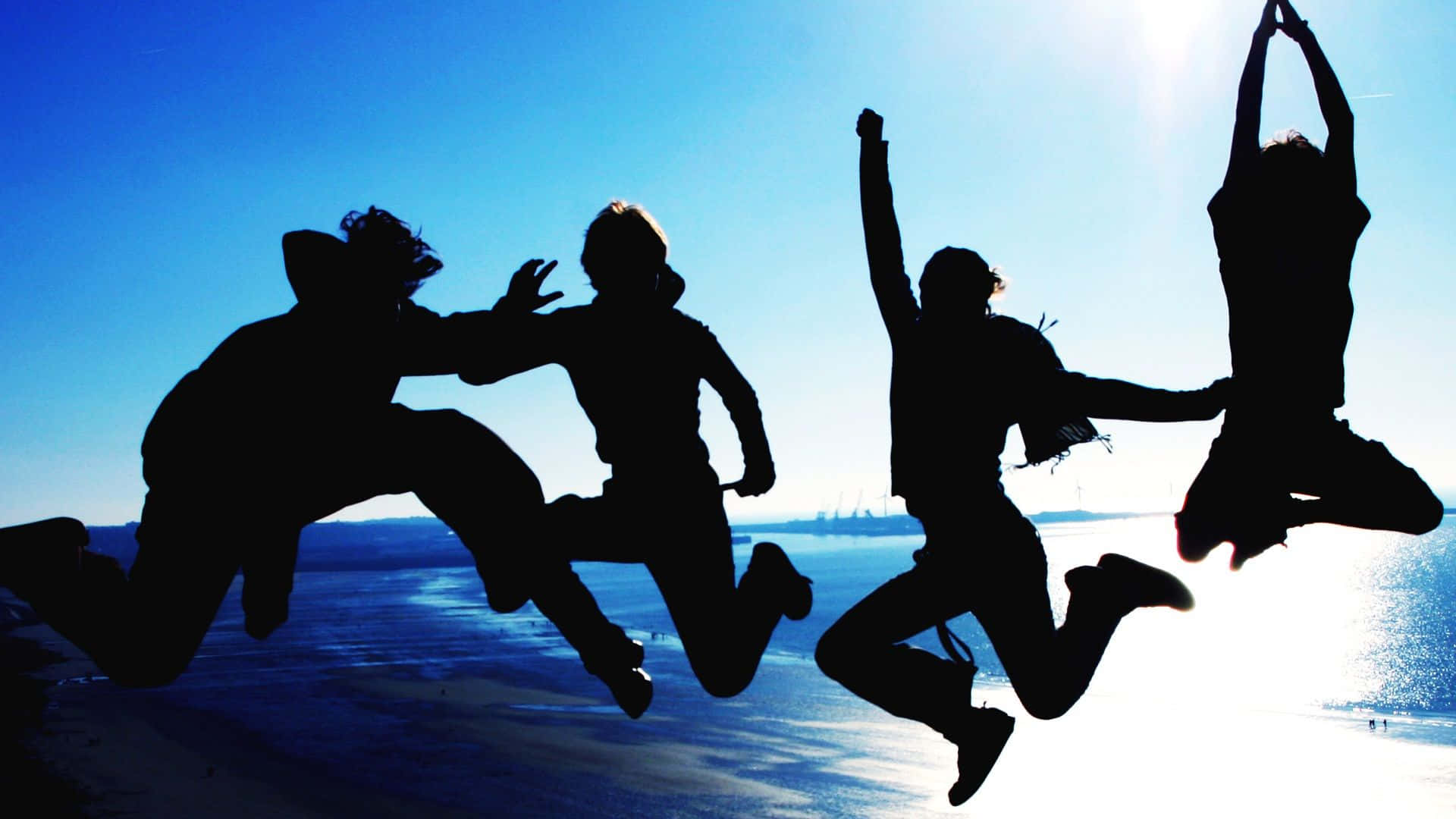 Friendship Day 桌布 壁紙 — 四個剪影朋友喺水上嘅明亮藍天下快樂咁跳起。