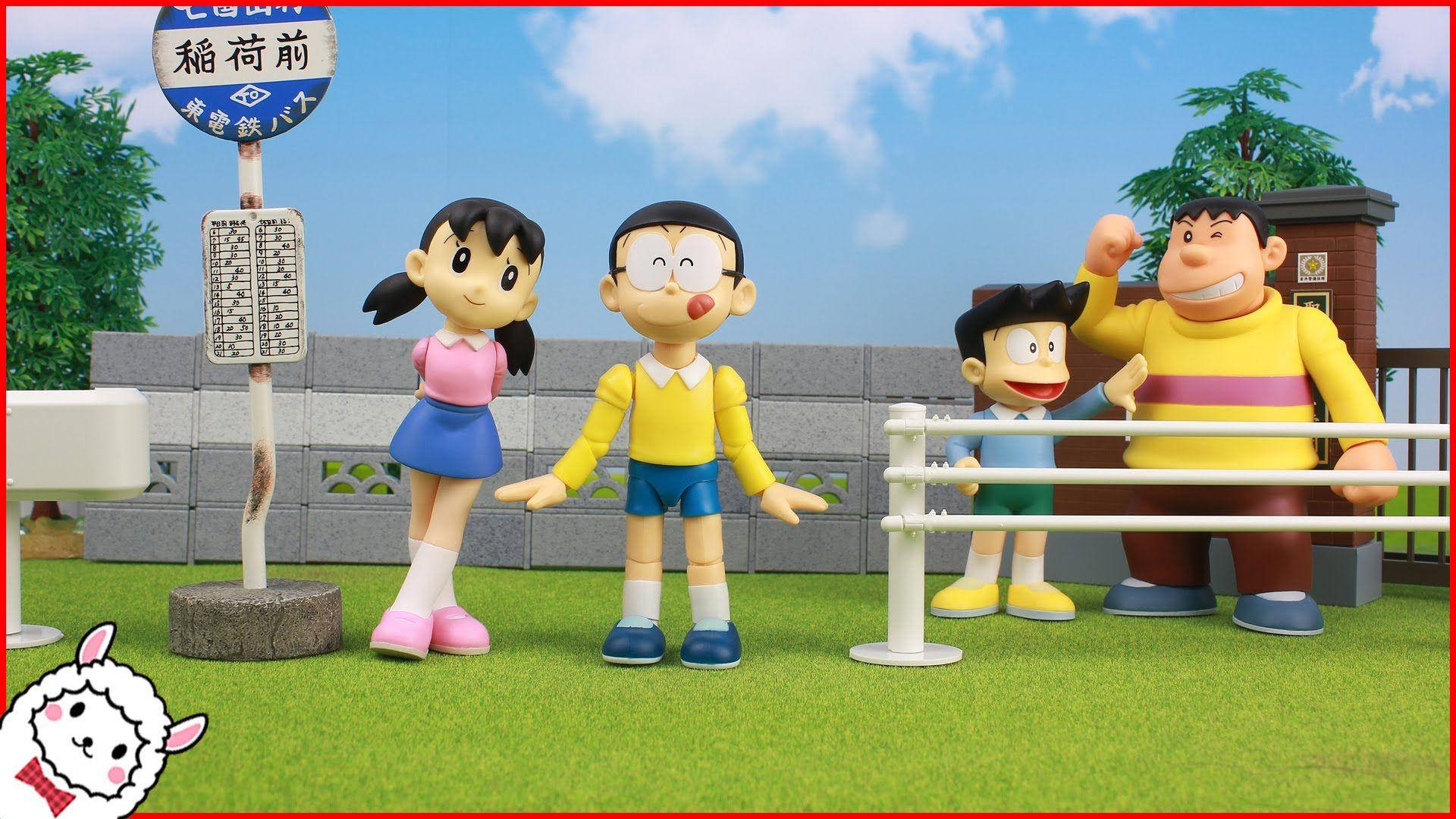 Doraemon 桌布 壁紙 — 四個多啦A夢角色站在草地上，靠近一個日本火車站標誌，背景是明亮的藍天。
