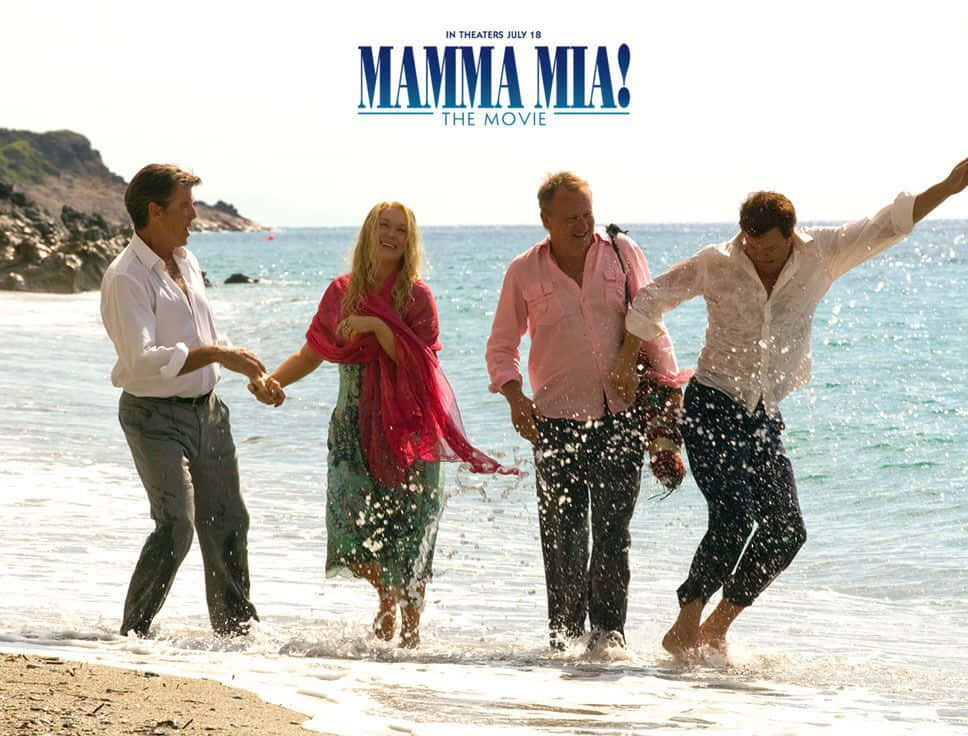 Mamma Mia 桌布 壁紙 — 四個快樂的人在陽光明媚的海灘上，背景是岩石峭壁，在淺海水中嬉戲濺水。