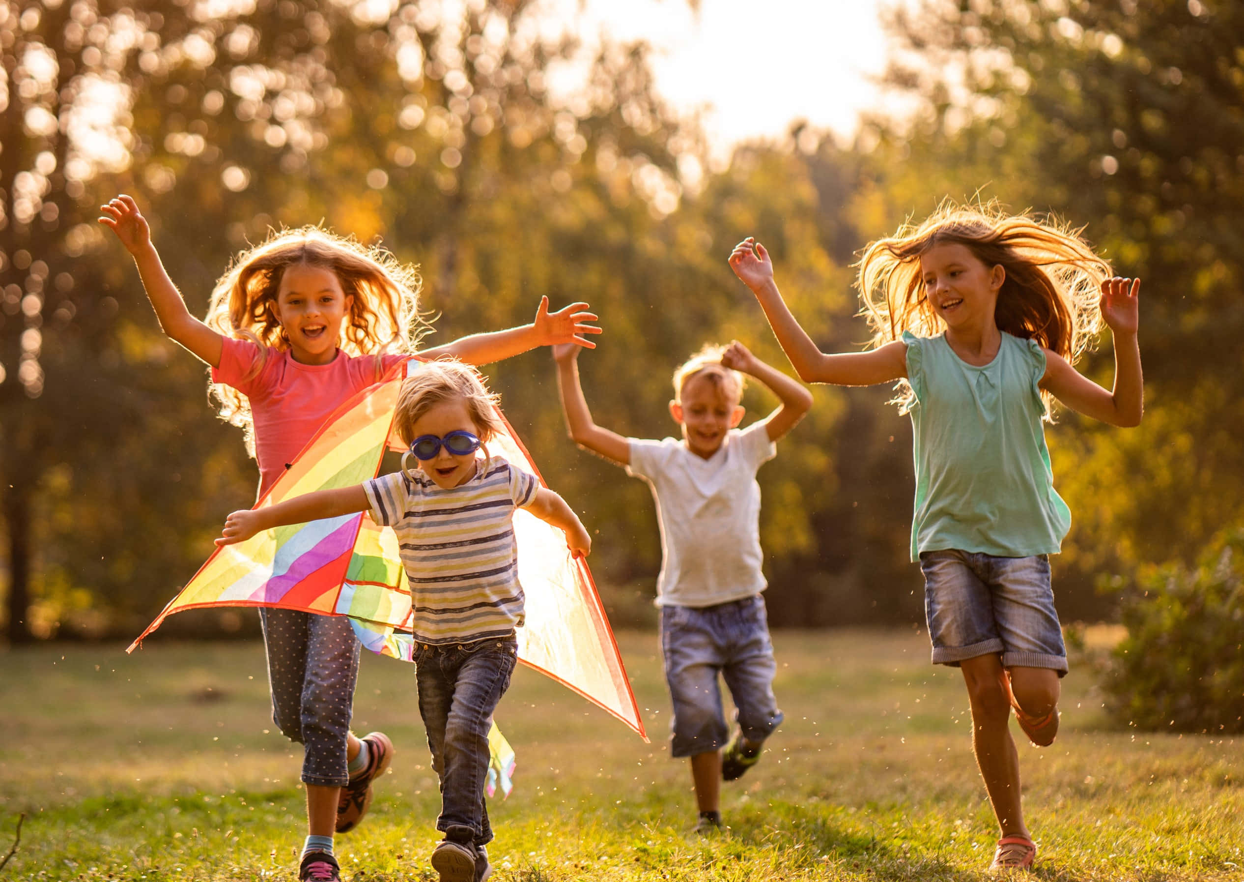 children 桌布 壁紙 — 四個快樂的小孩在陽光照耀的草地上奔跑，雙臂高舉，頭髮在金色的午後陽光中飛揚。