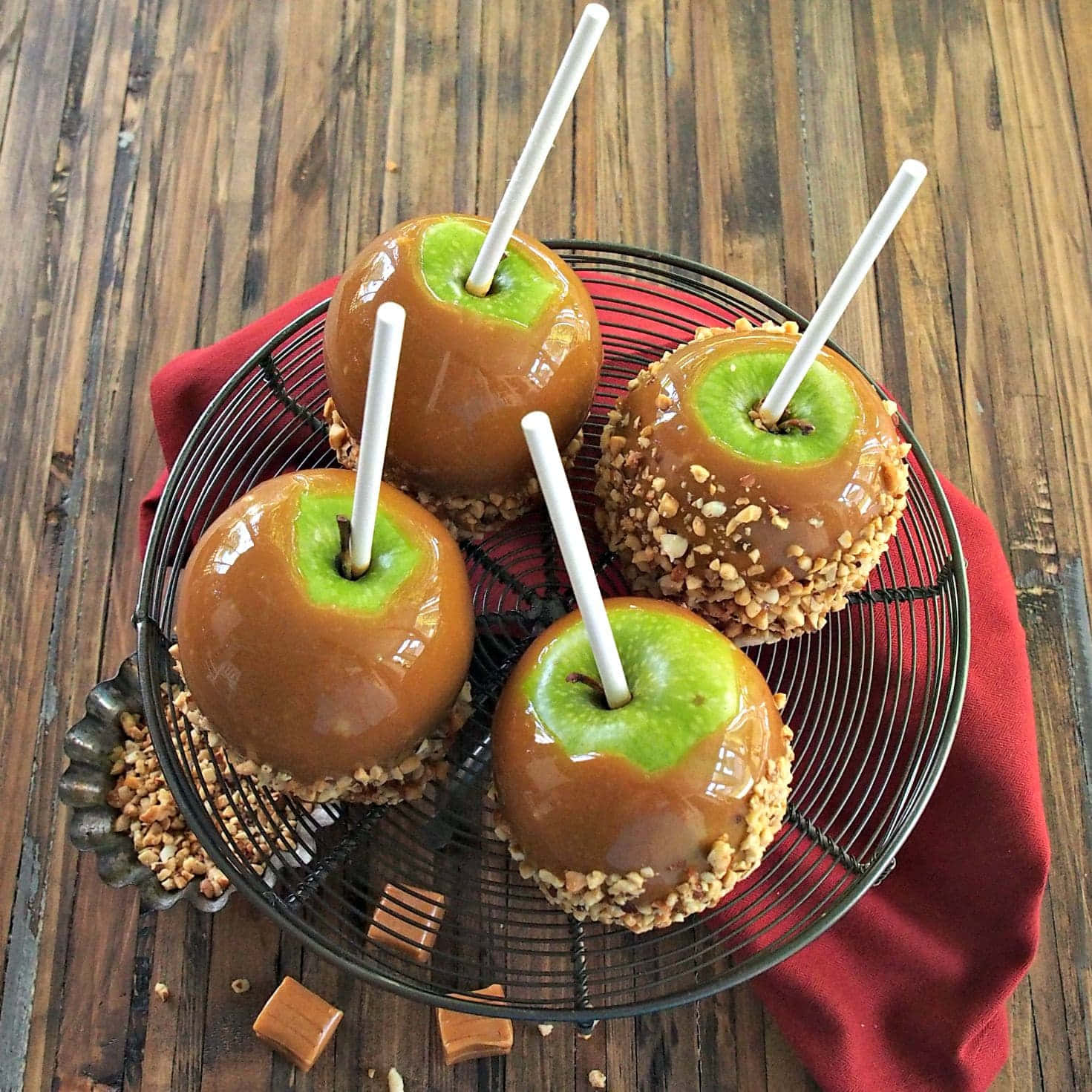 Caramel Apples 桌布 壁紙 — 四個焦糖包裹的蘋果，帶有綠色頂部和白色棍子，放在木質表面上的紅色布上的黑色金屬架上。