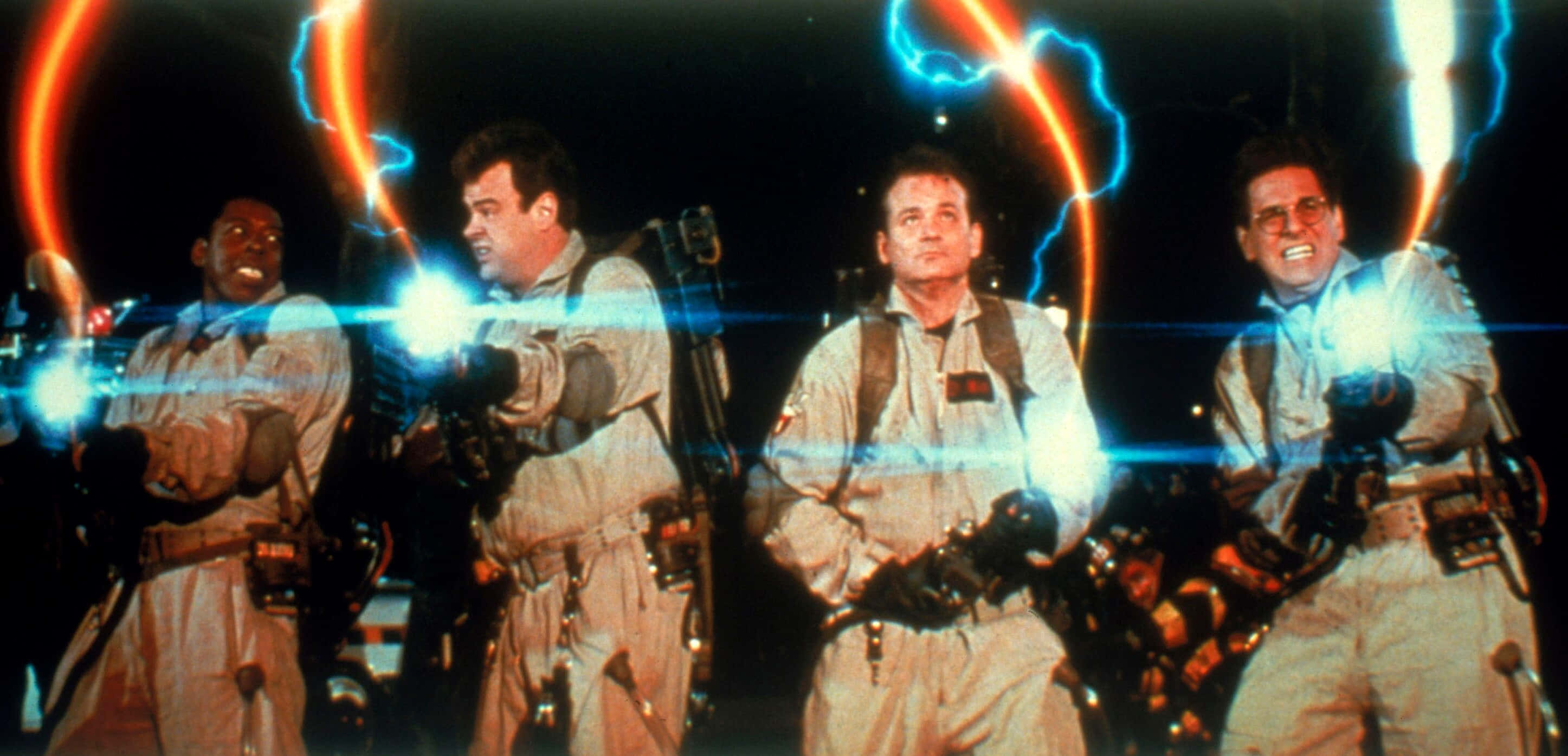 Ghostbusters 桌布 壁紙 — 四個穿著白色裝備的捉鬼敢死隊在黑暗背景下發射帶有發光藍色和橙色能量光束的質子背包。