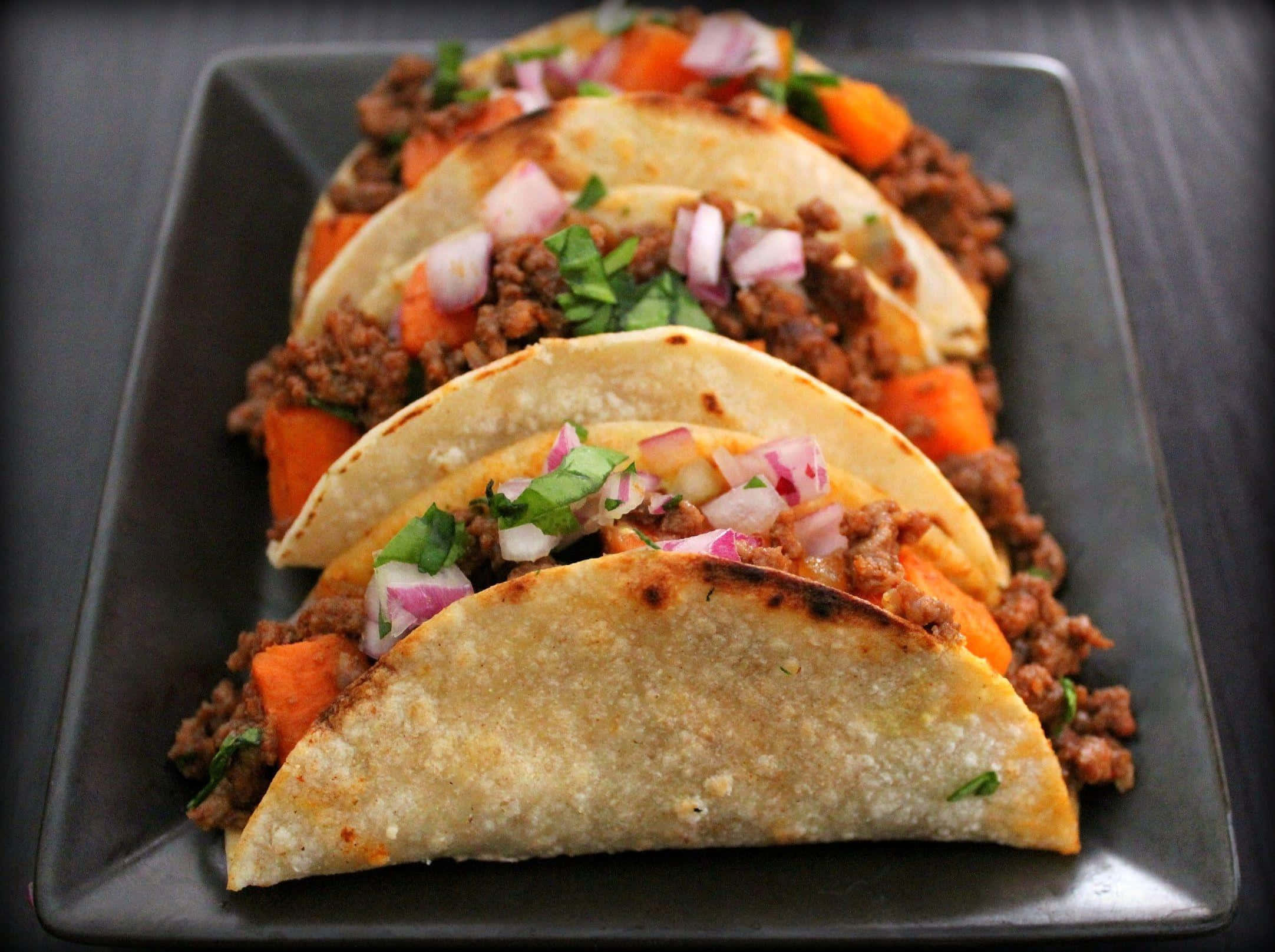 Taco 桌布 壁紙 — 四個脆皮墨西哥卷餅，內餡有調味肉、番薯和新鮮芫荽洋蔥，整齊地放在深色碟上。