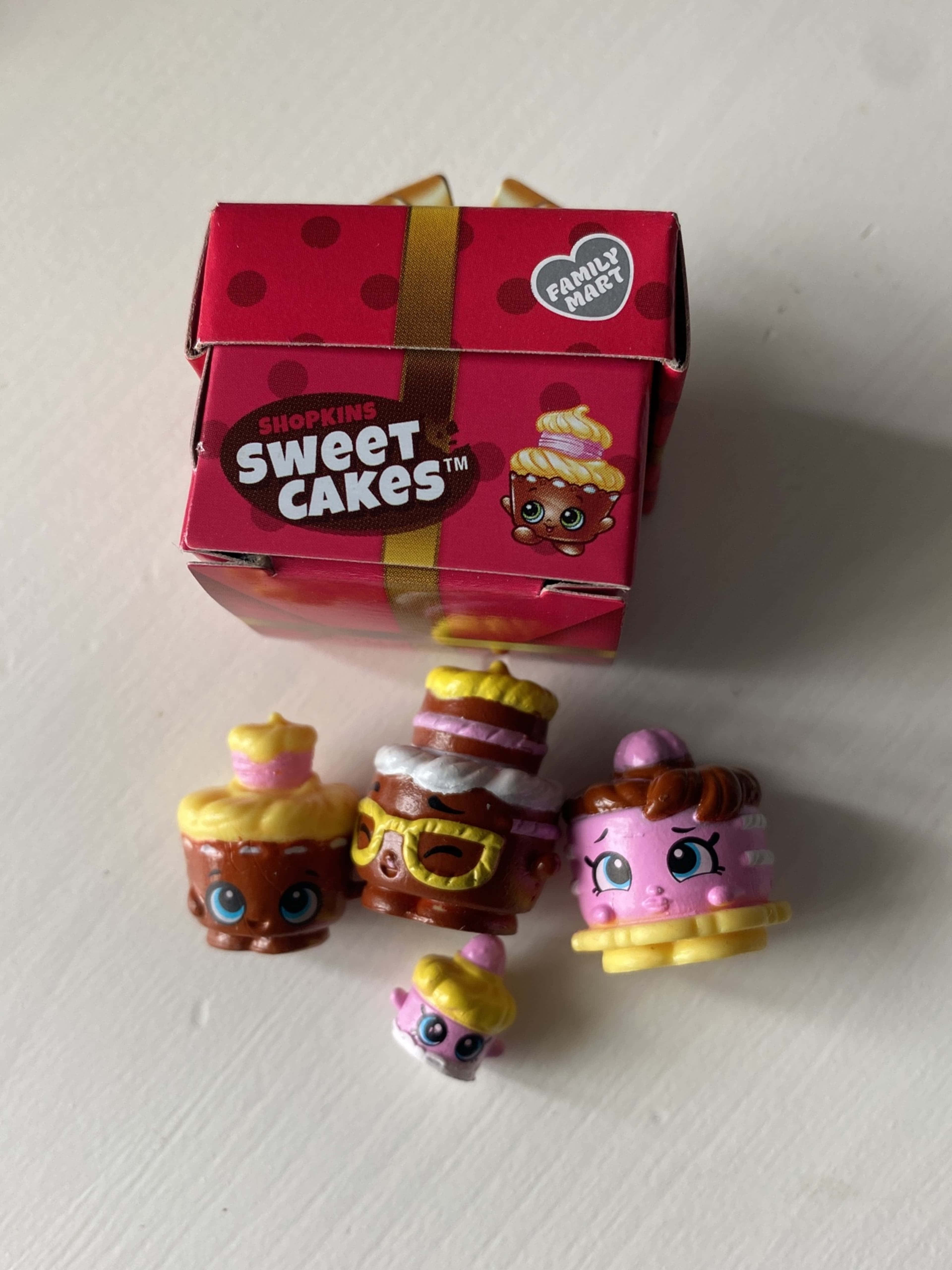 Shopkins 桌布 壁紙 — 四個色彩繽紛的Shopkins Sweet Cakes玩具放在白色表面上，旁邊是它們粉紅色圓點Family Mart盒子。