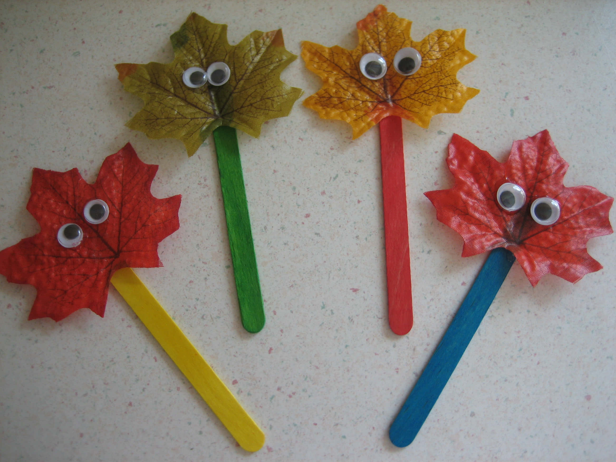Fall Crafts 桌布 壁紙 — 四個色彩繽紛的秋季樹葉手工藝品，帶有咕嚕眼睛貼在冰棒棍上，放在有斑點的檯面上。