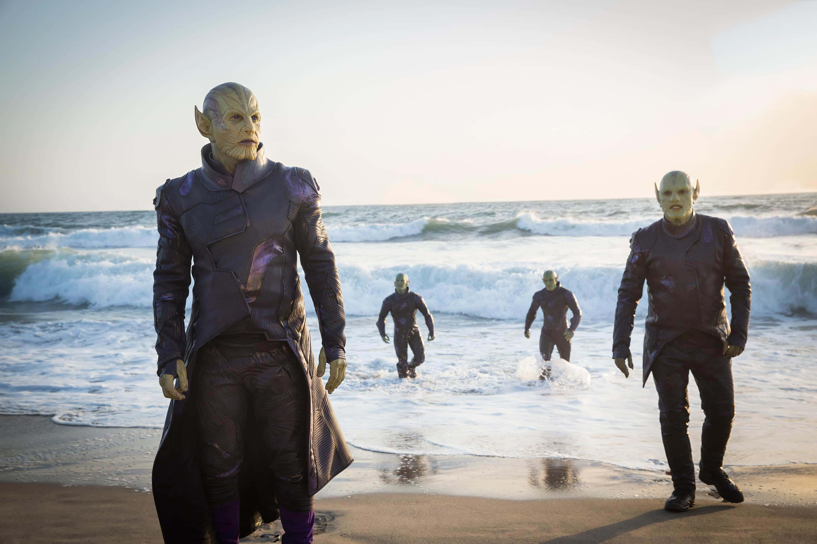 Skrulls 桌布 壁紙 — 四個身穿深色未來裝甲的外星人身影，沿著陽光明媚的海灘行走，身後是洶湧的海浪。