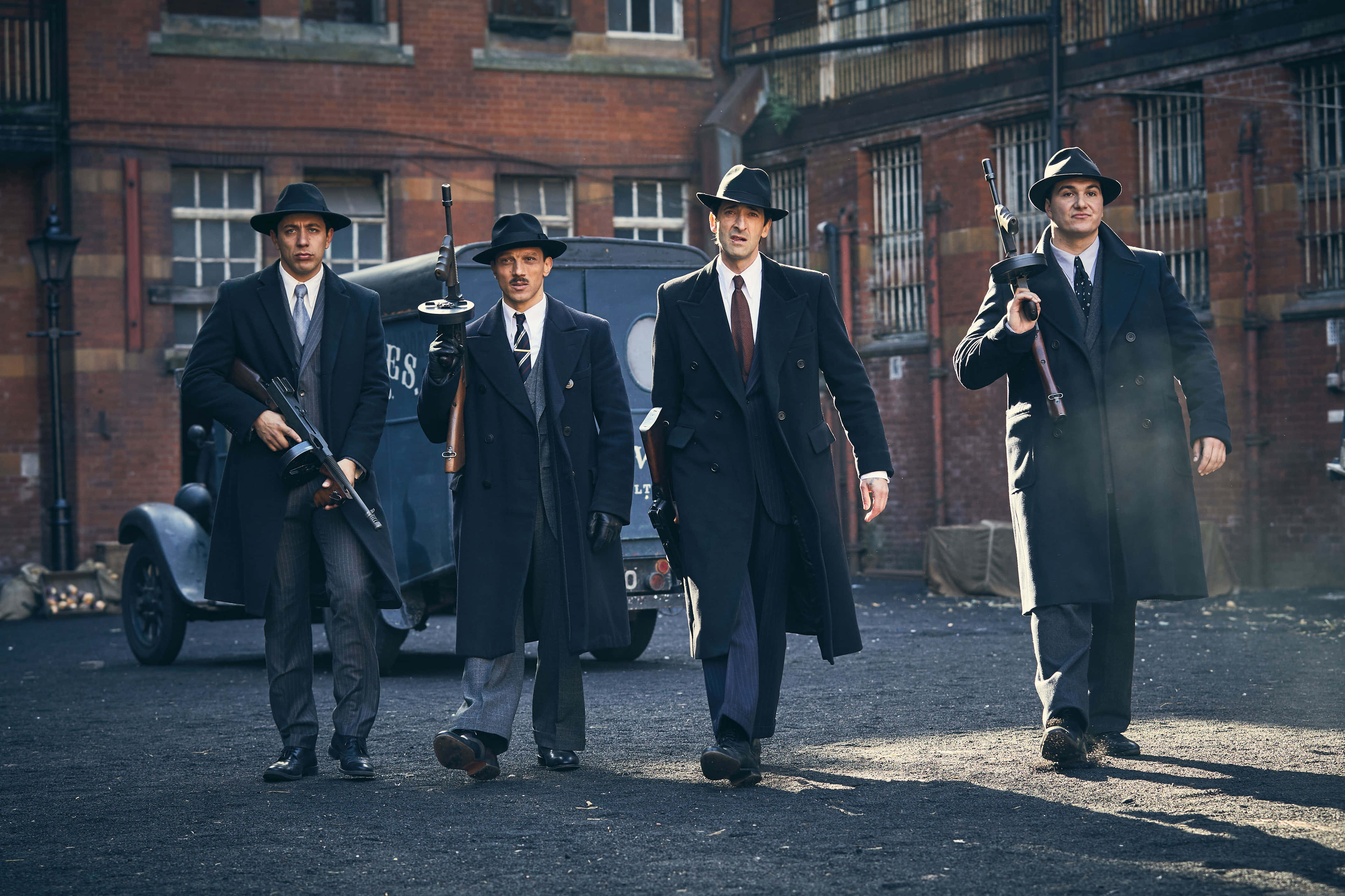 Peaky Blinders 桌布 壁紙 — 四個身穿深色西裝和軟呢帽的男人，在背景有復古汽車和磚砌建築的骯髒城市街道上，有目的地行走。