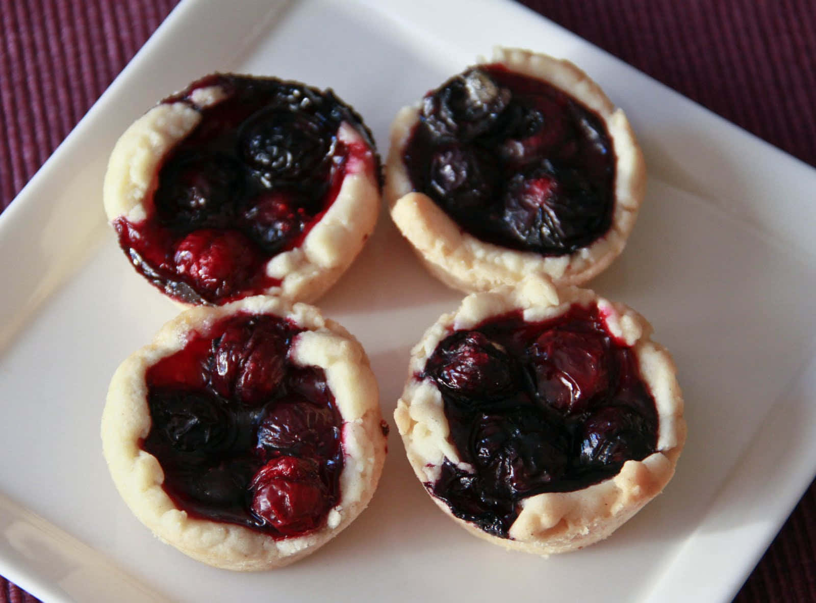 Blueberries Tart 桌布 壁紙 — 四個迷你莓果撻，帶有光澤的深色水果餡料，放在白色方形碟上，背景為深紫色紋理。