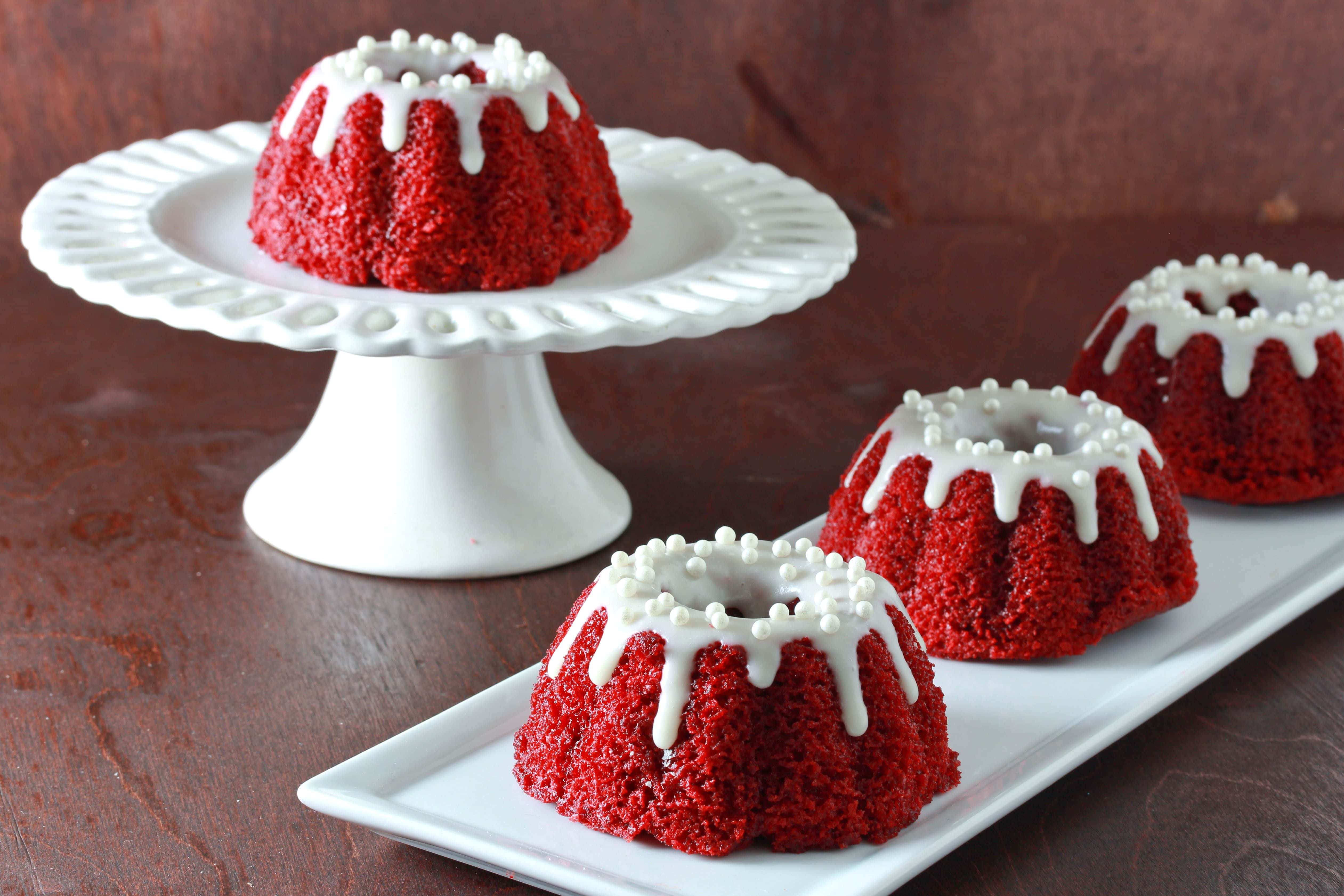 Red Velvet Cake 桌布 壁紙 — 四個鮮紅色的紅絲絨圓環蛋糕，配上白色糖霜和珍珠糖粒，優雅地擺放在白色陶瓷碟上，背景是質樸的木質背景。