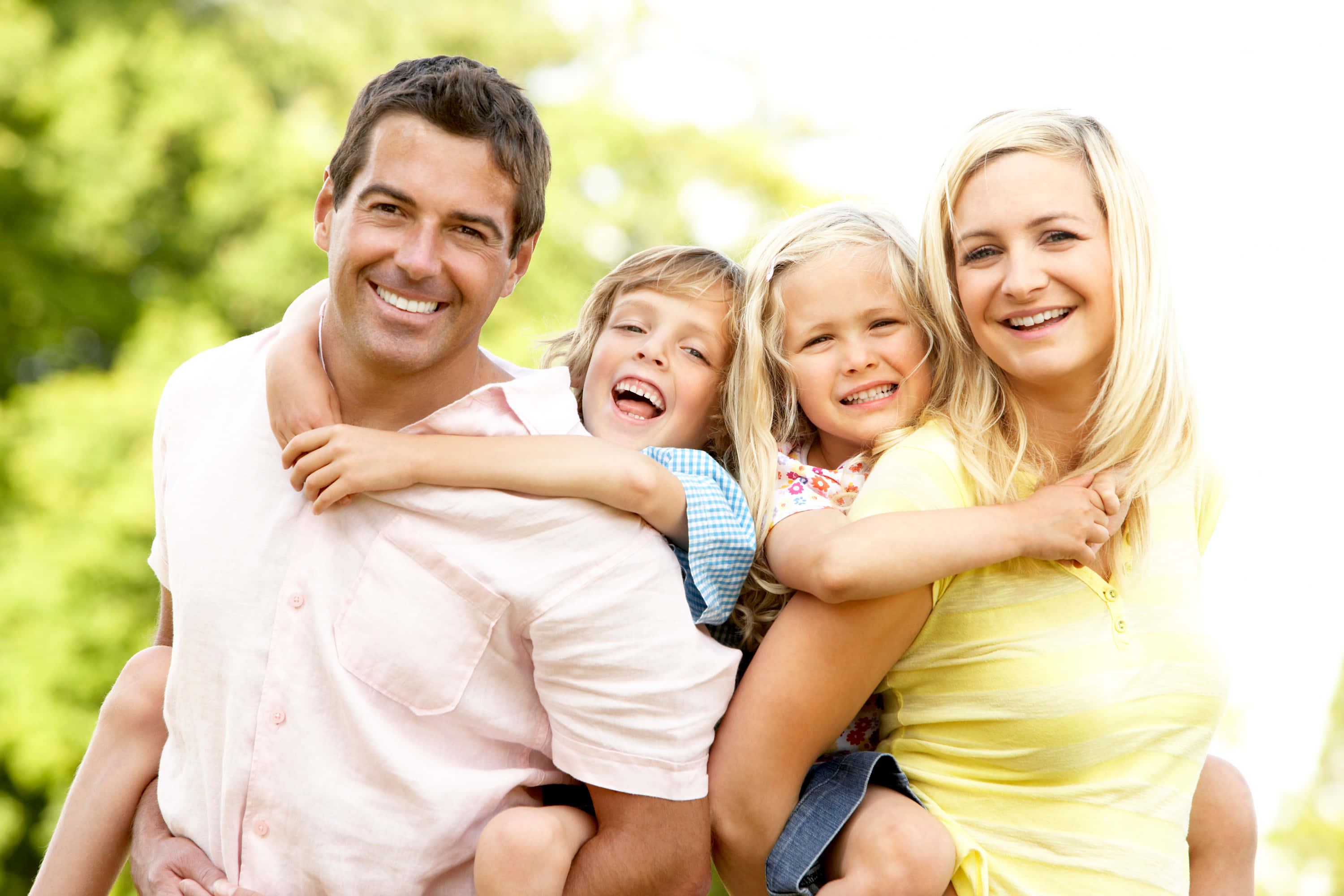 四口之家慶祝生活 壁紙 — 一個快樂的四口之家在戶外燦爛地微笑，背後有柔和的綠色樹葉模糊效果。
