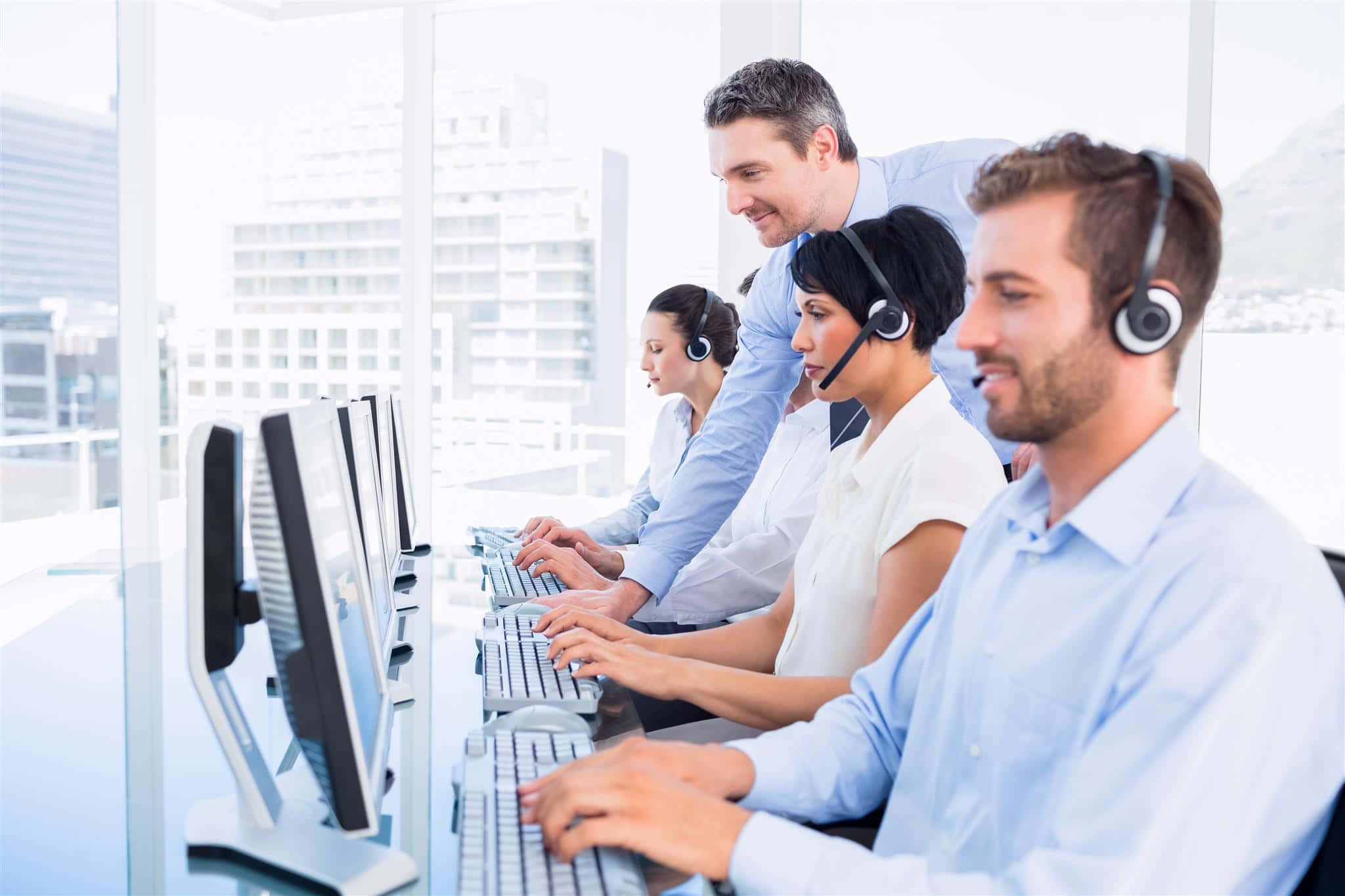 Telemarketing 桌布 壁紙 — 四名穿著淺藍色襯衫和耳機的客戶服務人員在一個明亮現代、可欣賞城市景觀的辦公室裡使用電腦工作。