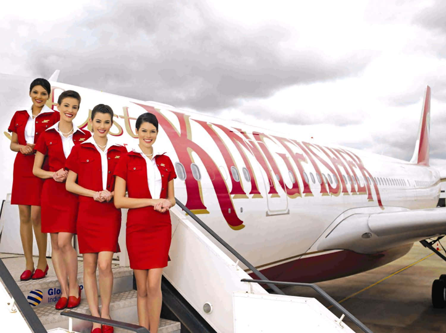 Flight Attendant 桌布 壁紙 — 四名身穿紅色制服、面帶微笑的空中服務員站在翠鳥航空飛機的樓梯上，天空多雲。