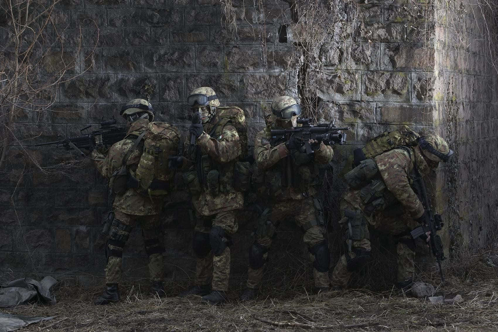 Army 桌布 壁紙 — 四名身穿迷彩裝備的士兵蹲在風化的石牆後面，手持步槍，處於緊張的戰鬥姿態。