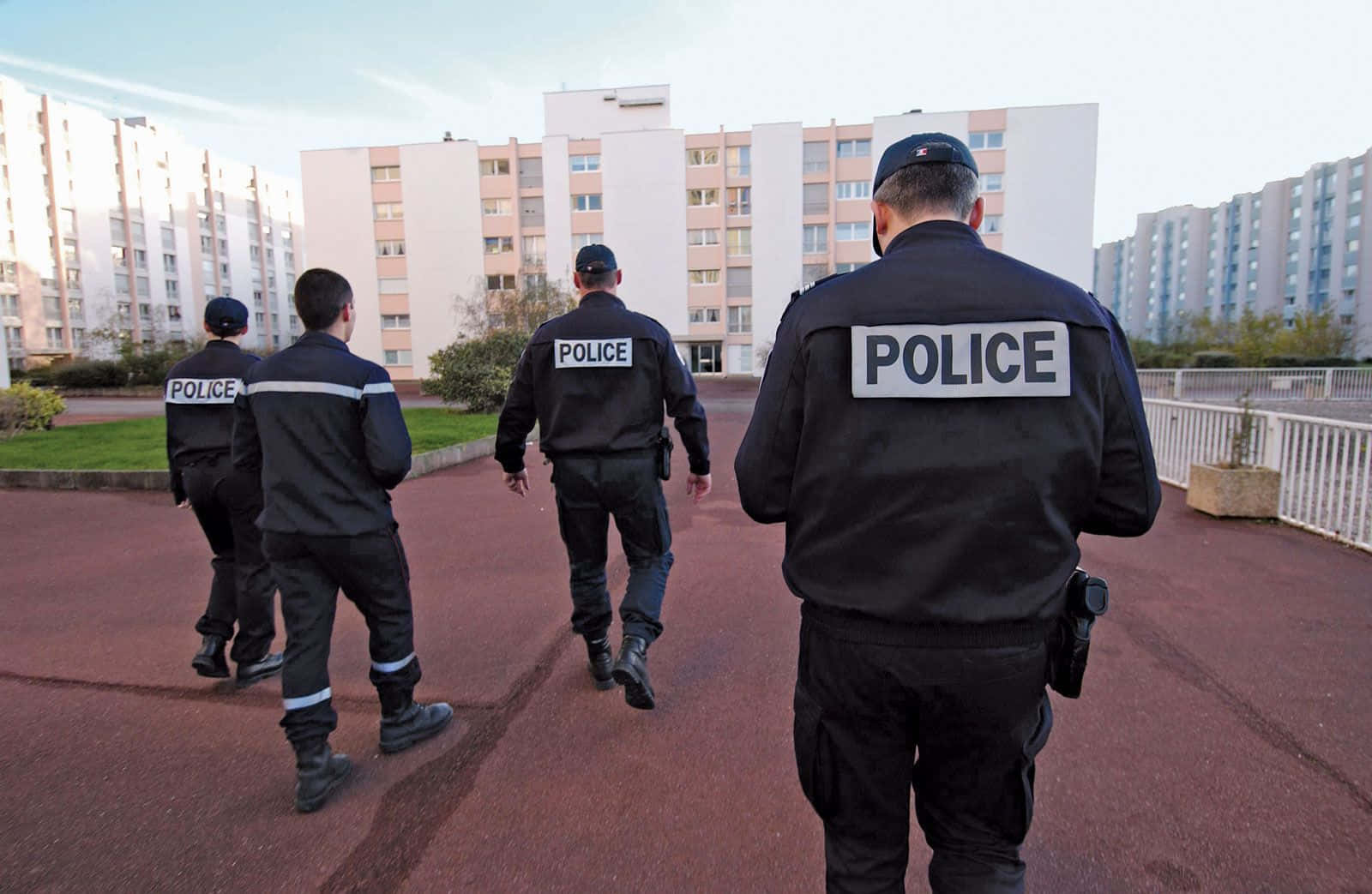 警察 桌布 壁紙 — 四名身穿黑色制服的警察沿著一條紅色小徑遠離鏡頭，走向蒼白天空下的公寓大樓。