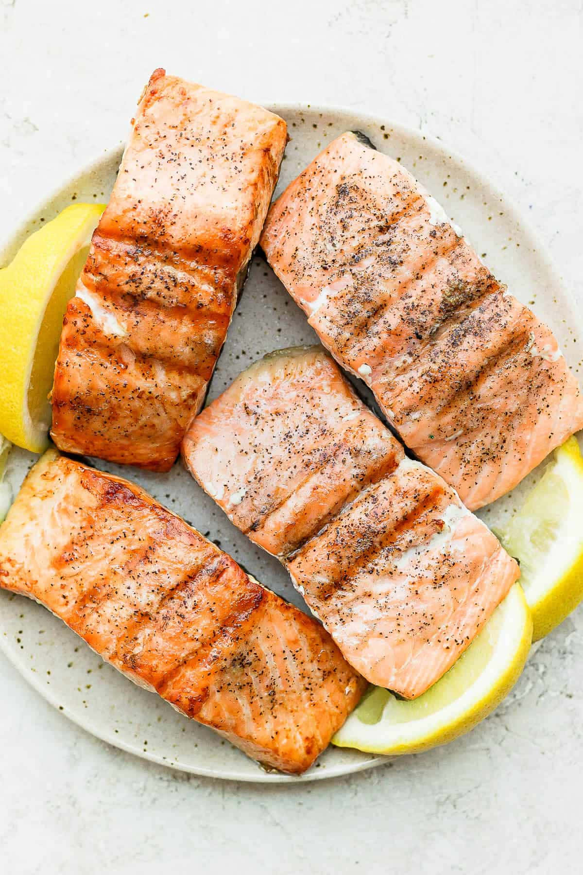 Salmon 桌布 壁紙 — 四塊帶有烤痕和黑胡椒的烤三文魚柳放在有斑點的碟上，旁邊有檸檬角。