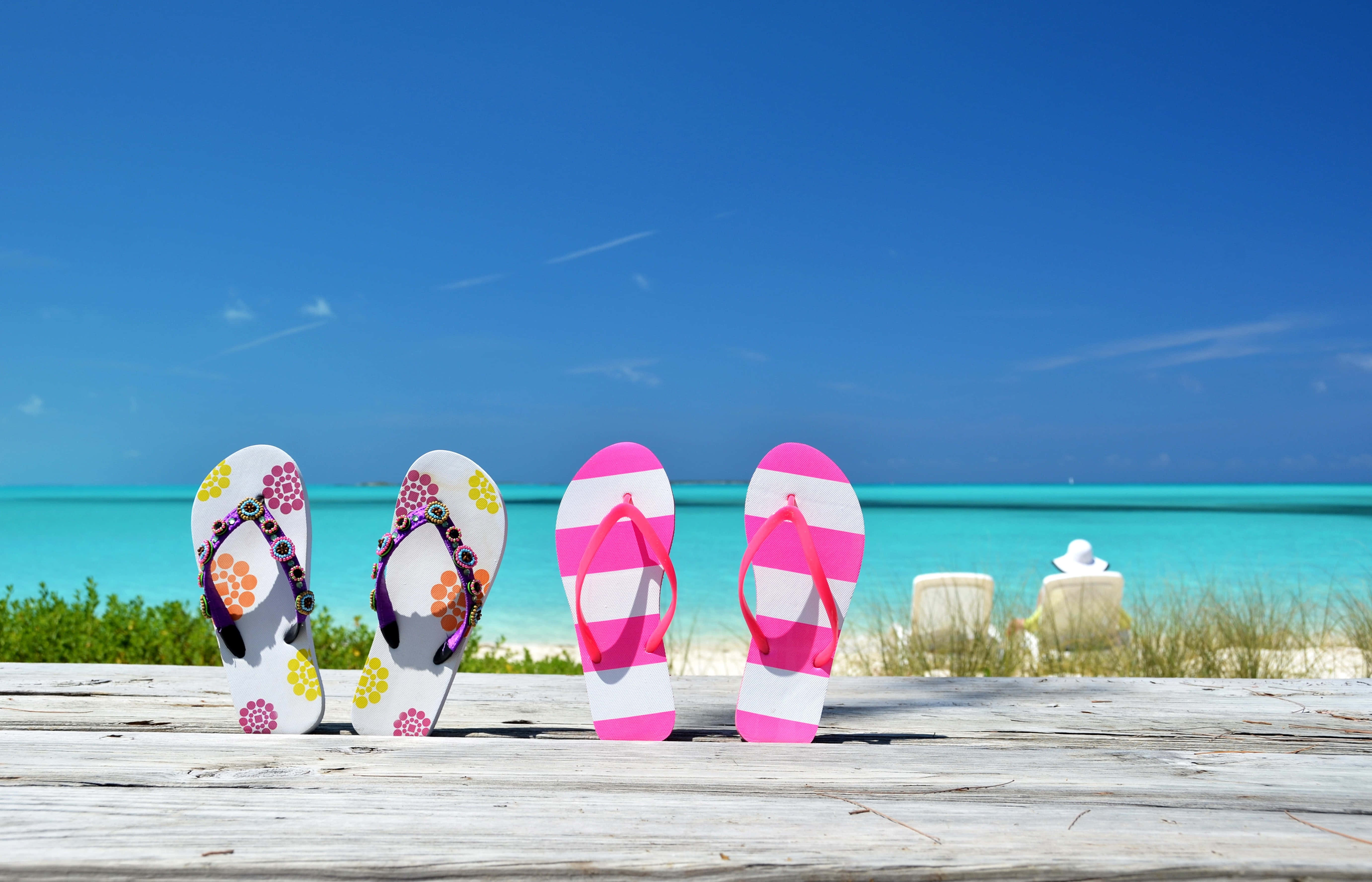 Beach Sandals 桌布 壁紙 — 四對色彩繽紛的拖鞋放在一個飽經風霜的木甲板上，俯瞰著清澈藍天下的碧綠海洋。