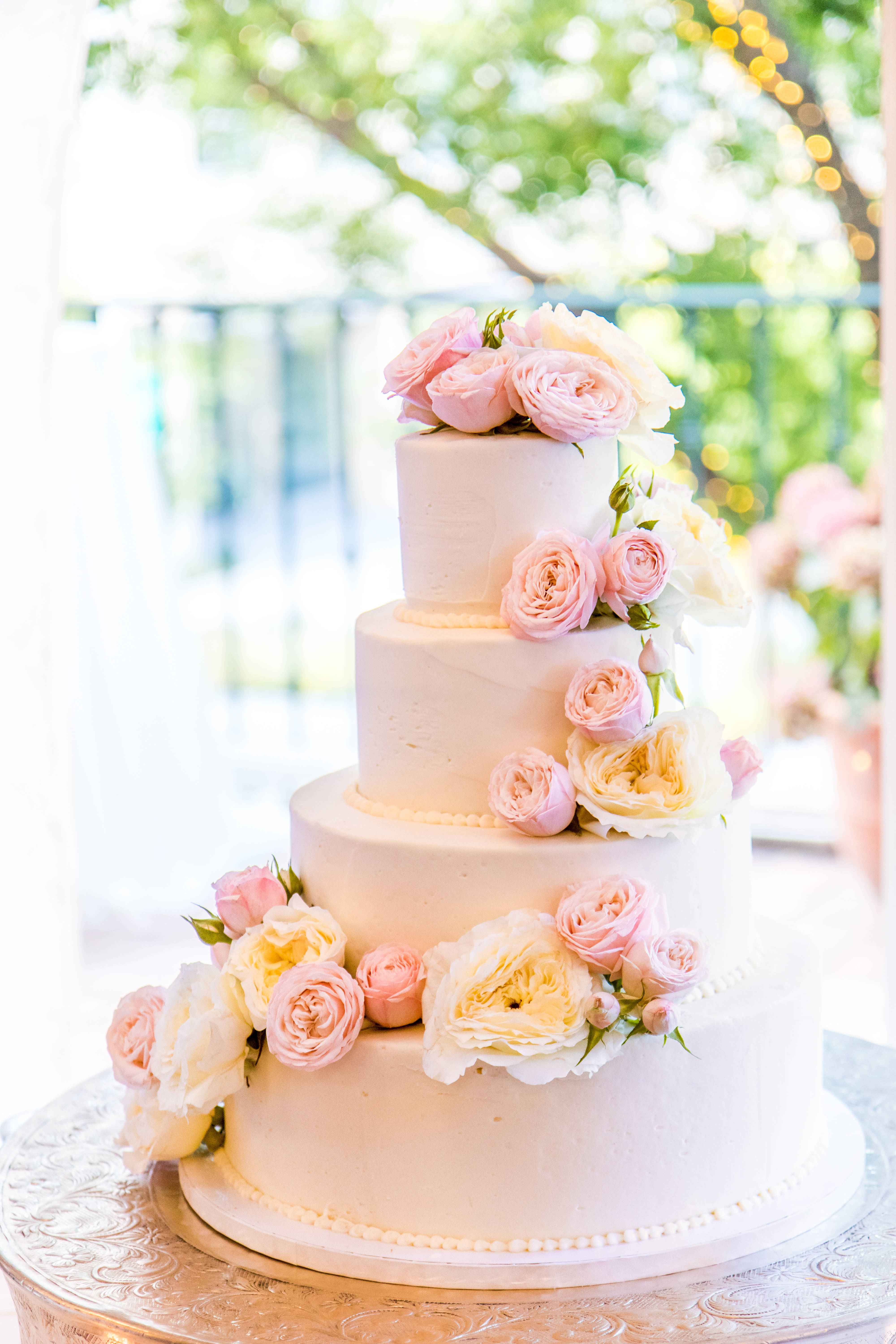 四層圓形花卉婚禮蛋糕 壁紙 — 一個四層白色結婚蛋糕，飾以柔和粉紅和奶油色玫瑰，優雅地放在銀色架上，在明亮的自然光下。