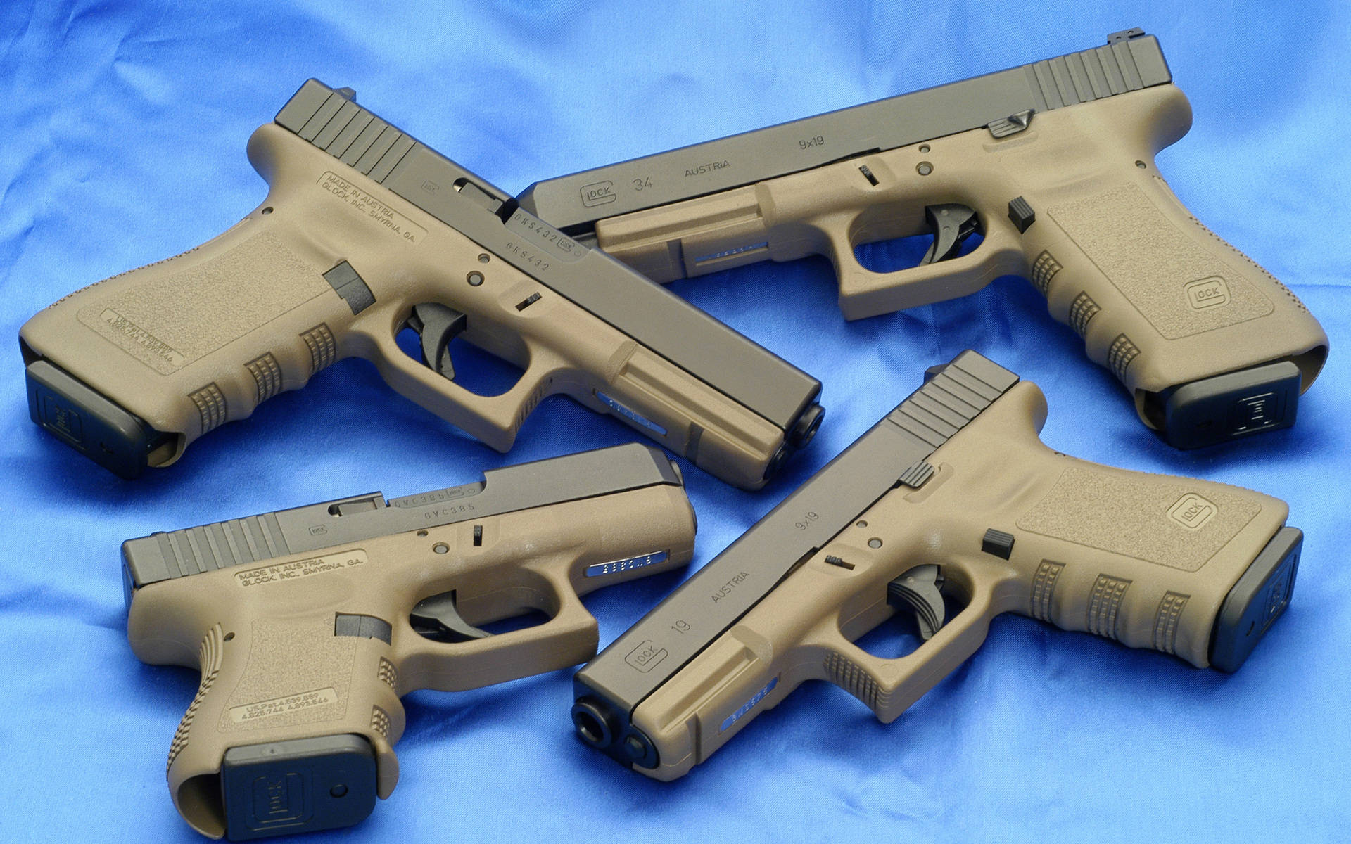 四把軍綠色格洛克壁紙 壁紙 — 四支淺褐色 Glock 手槍排列在帶紋理的藍色布料背景上，呈現產品風格照片。
